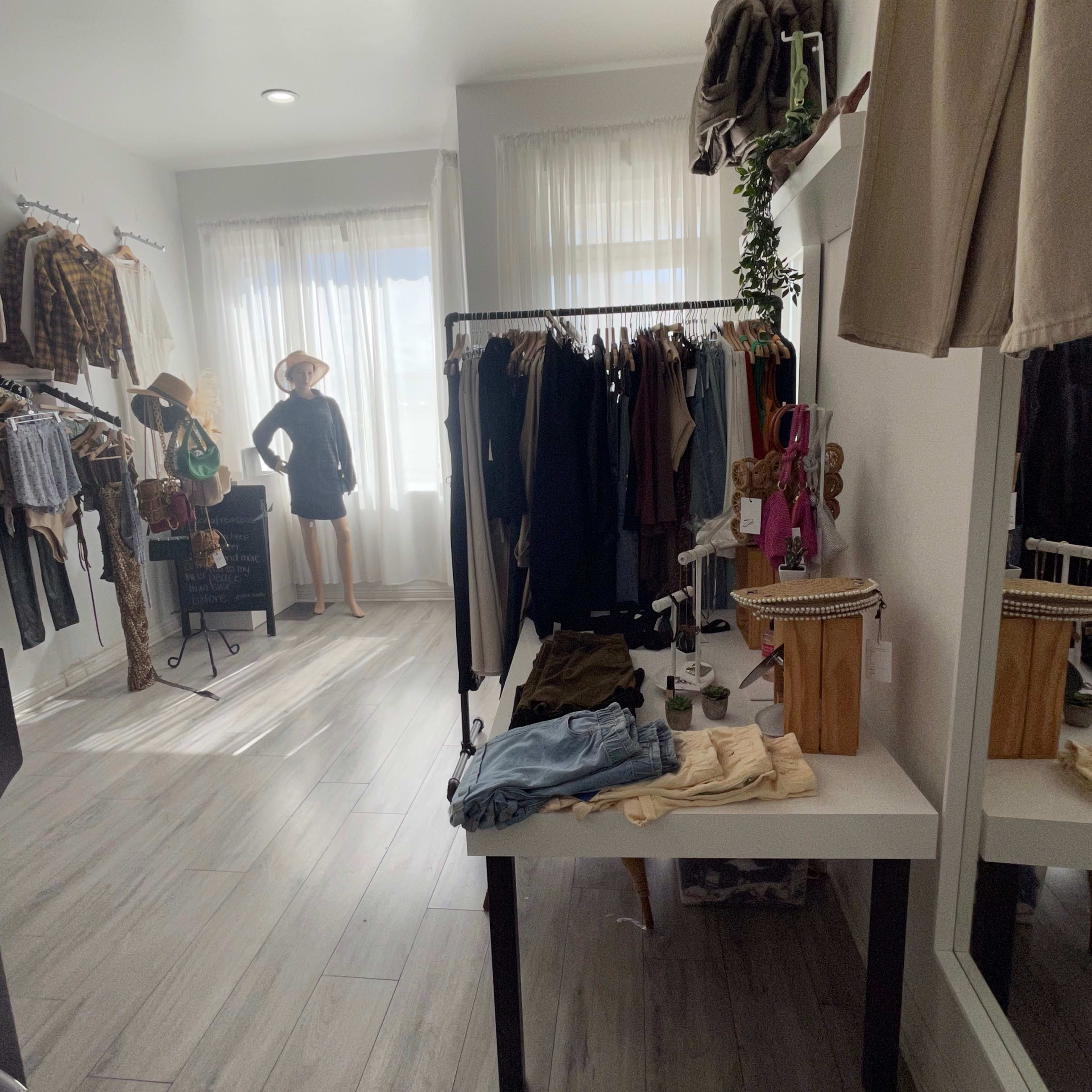 A boutique interior with clothing racks on the walls, a display table with folded garments, and a mannequin dressed in casual clothing near a window.