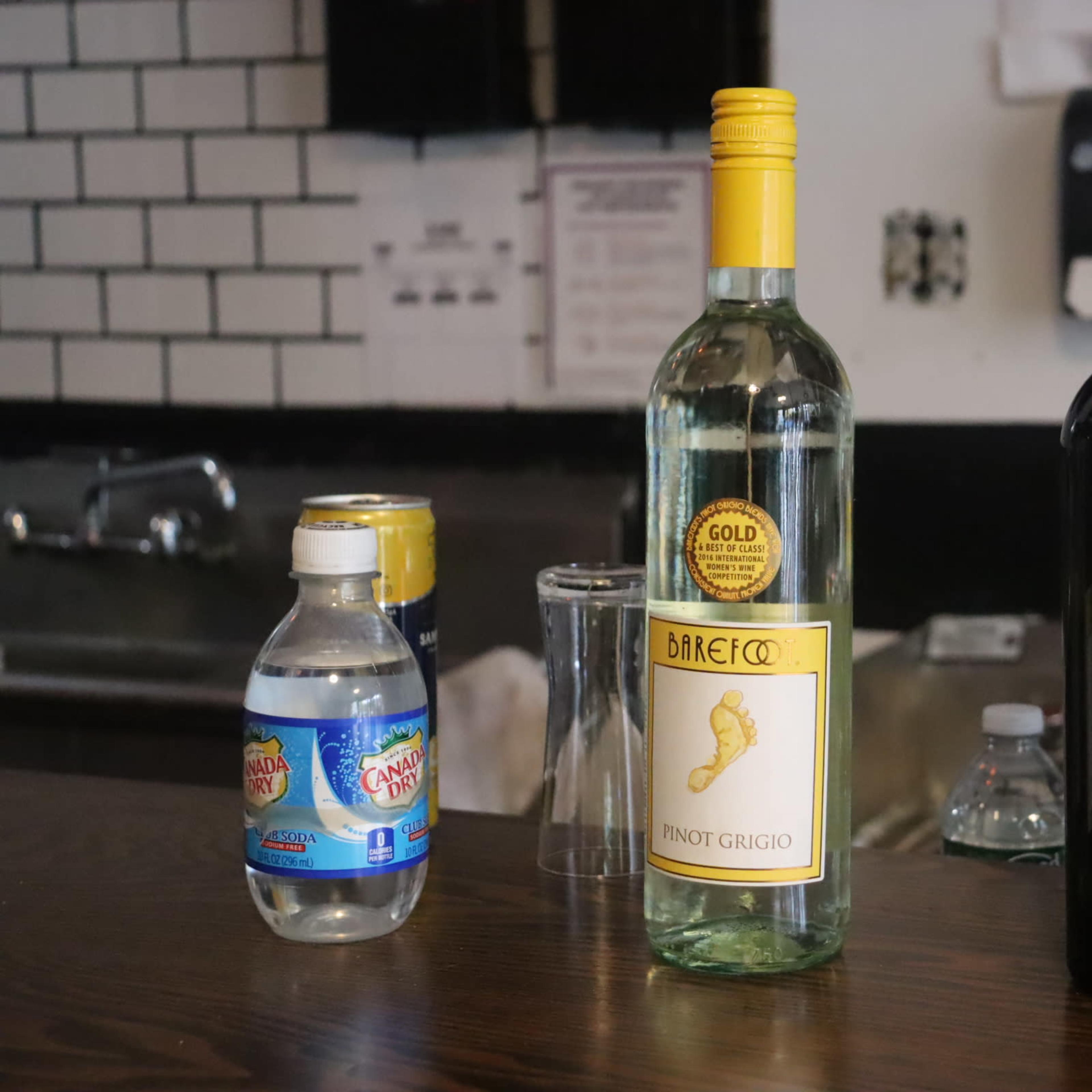A bar countertop with a bottle of Pinot Grigio, a bottle of red wine labeled "ombu," a can of soda, a plastic water bottle, and a glass.