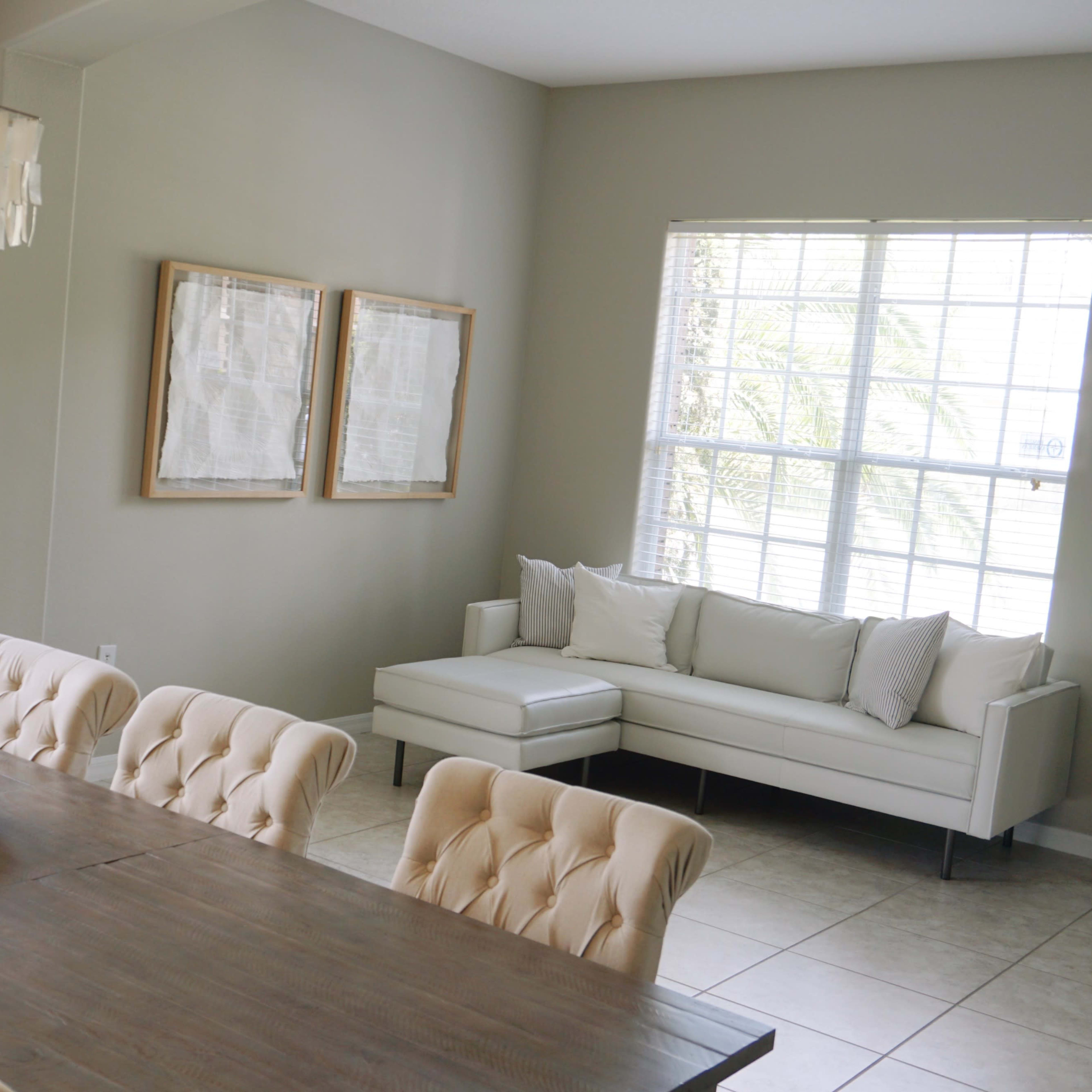 A dining area features a long wooden table with tufted chairs, a chandelier above, and a light-filled corner with a white sofa and framed artwork on the walls.