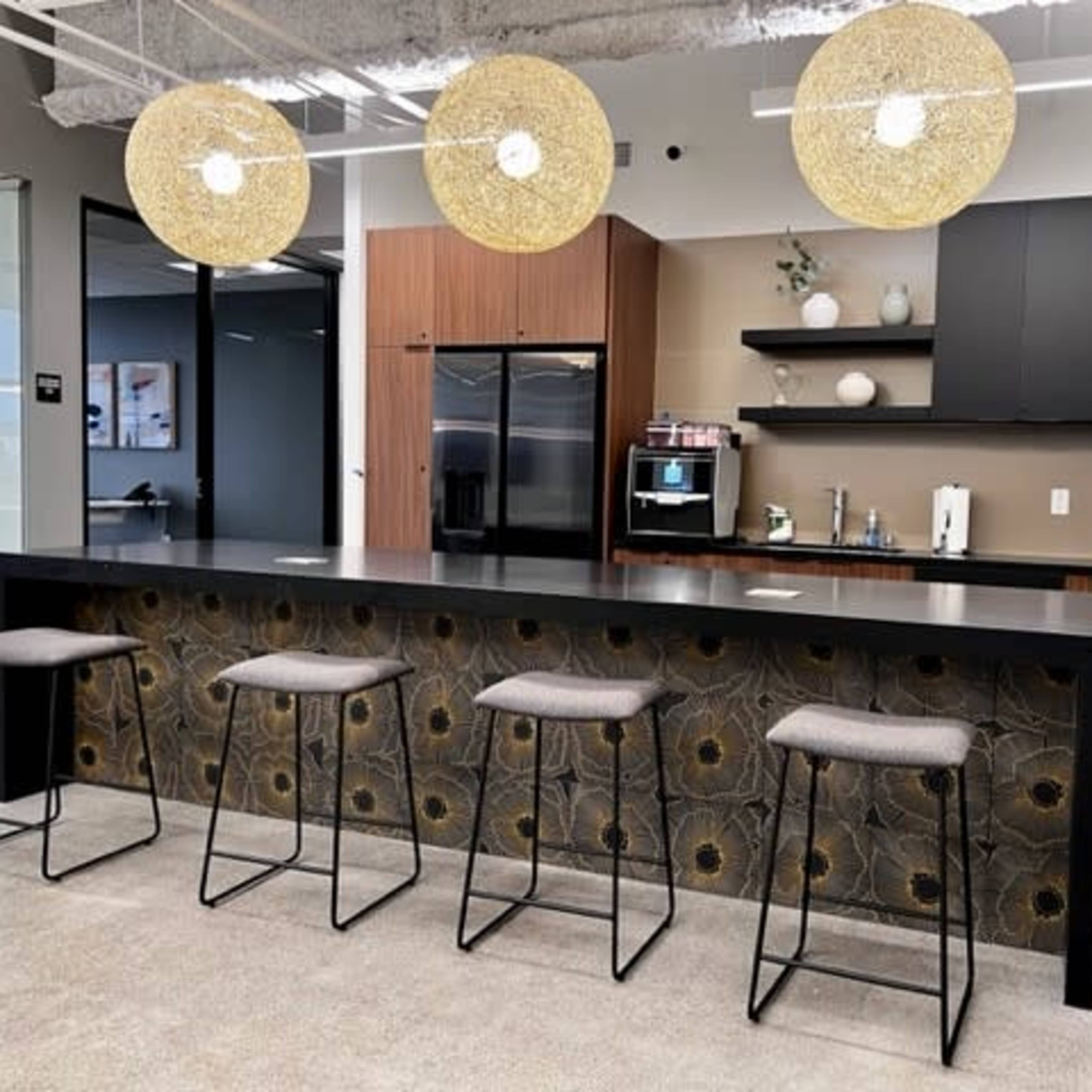 A modern kitchen area featuring a dark countertop, a row of gray bar stools, and unique pendant lights hanging above.