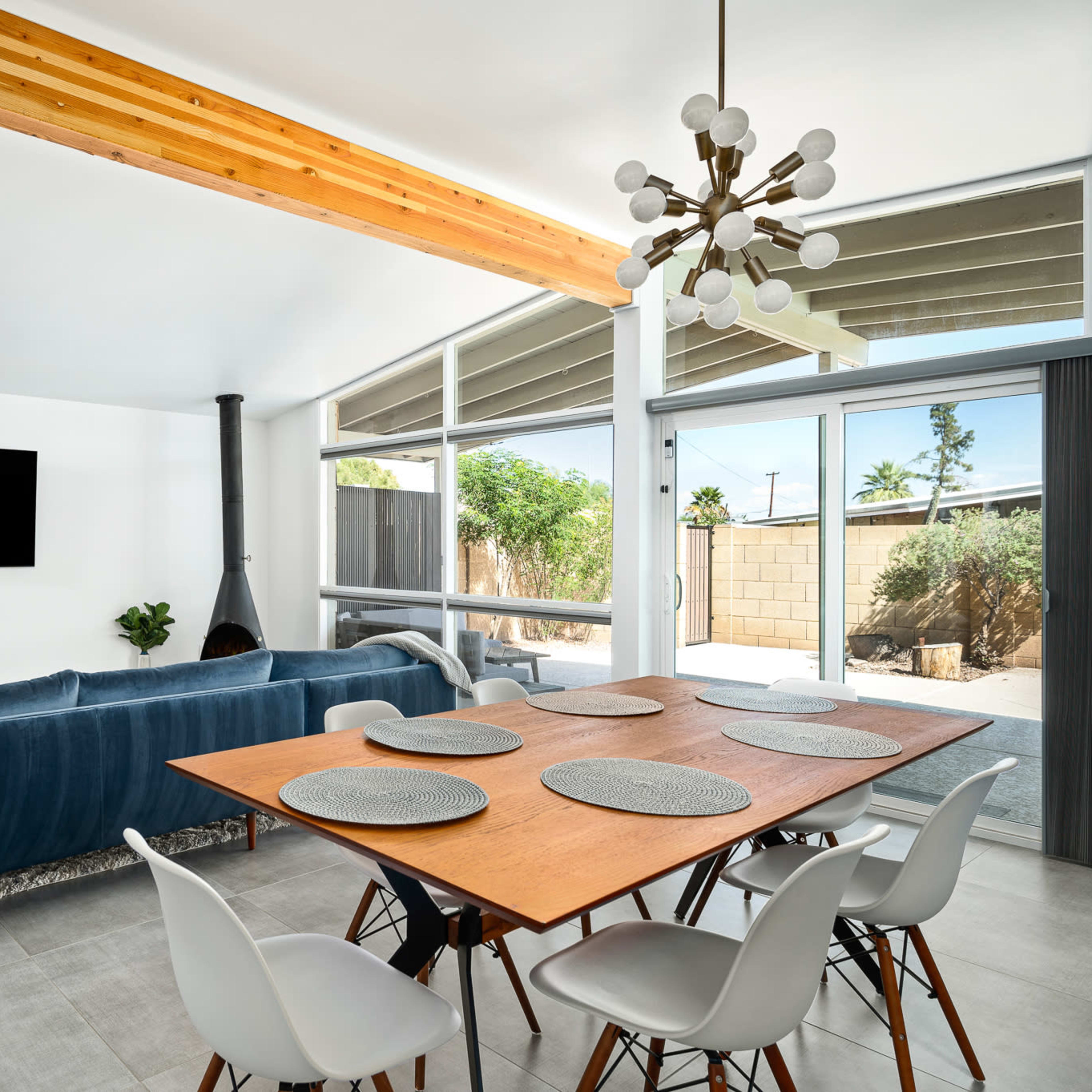 A modern living area features a blue sofa, a wooden dining table set for six, and large windows with a view of a patio garden.
