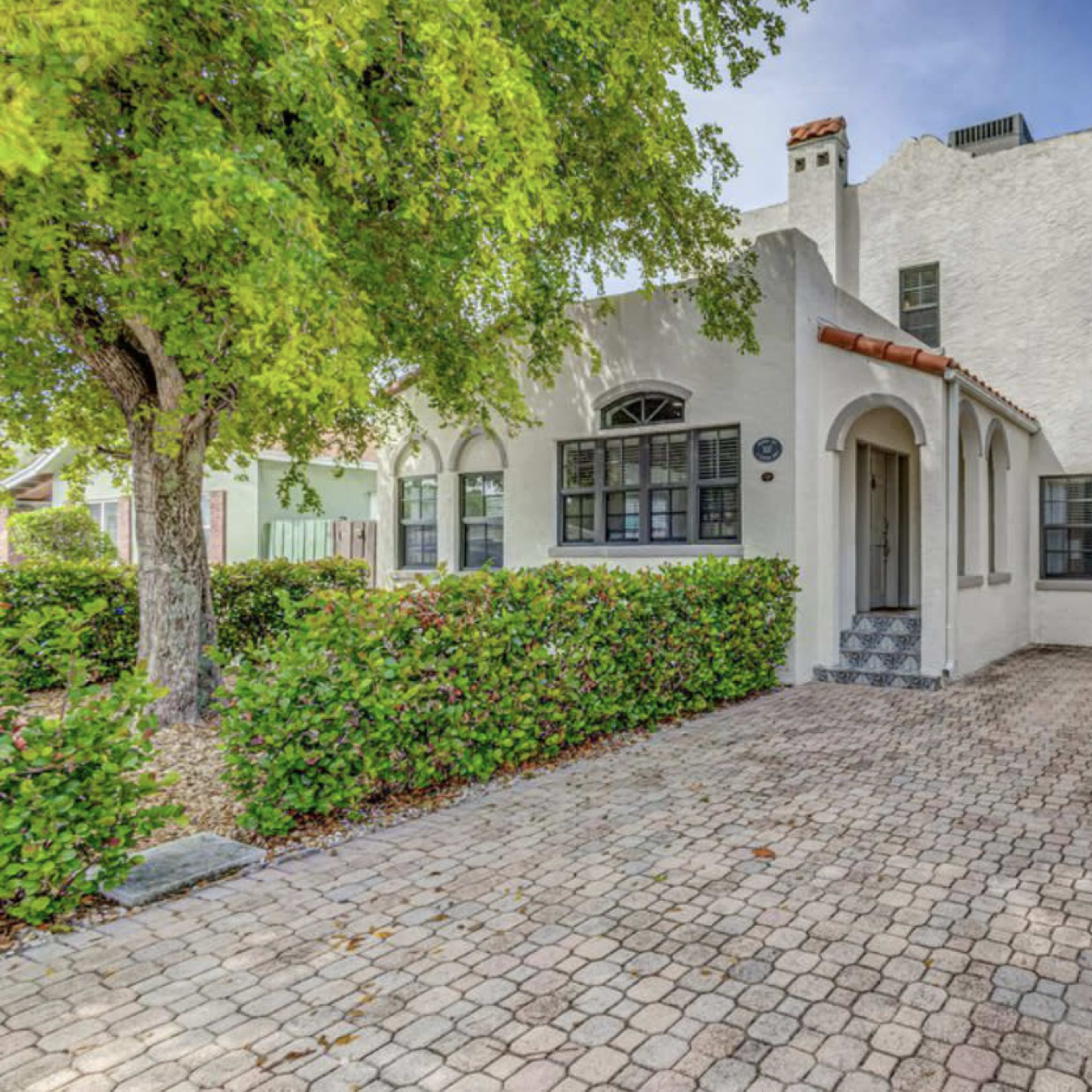 A white, two-story house with a tiled roof and a cobblestone driveway surrounded by green shrubs and trees.
