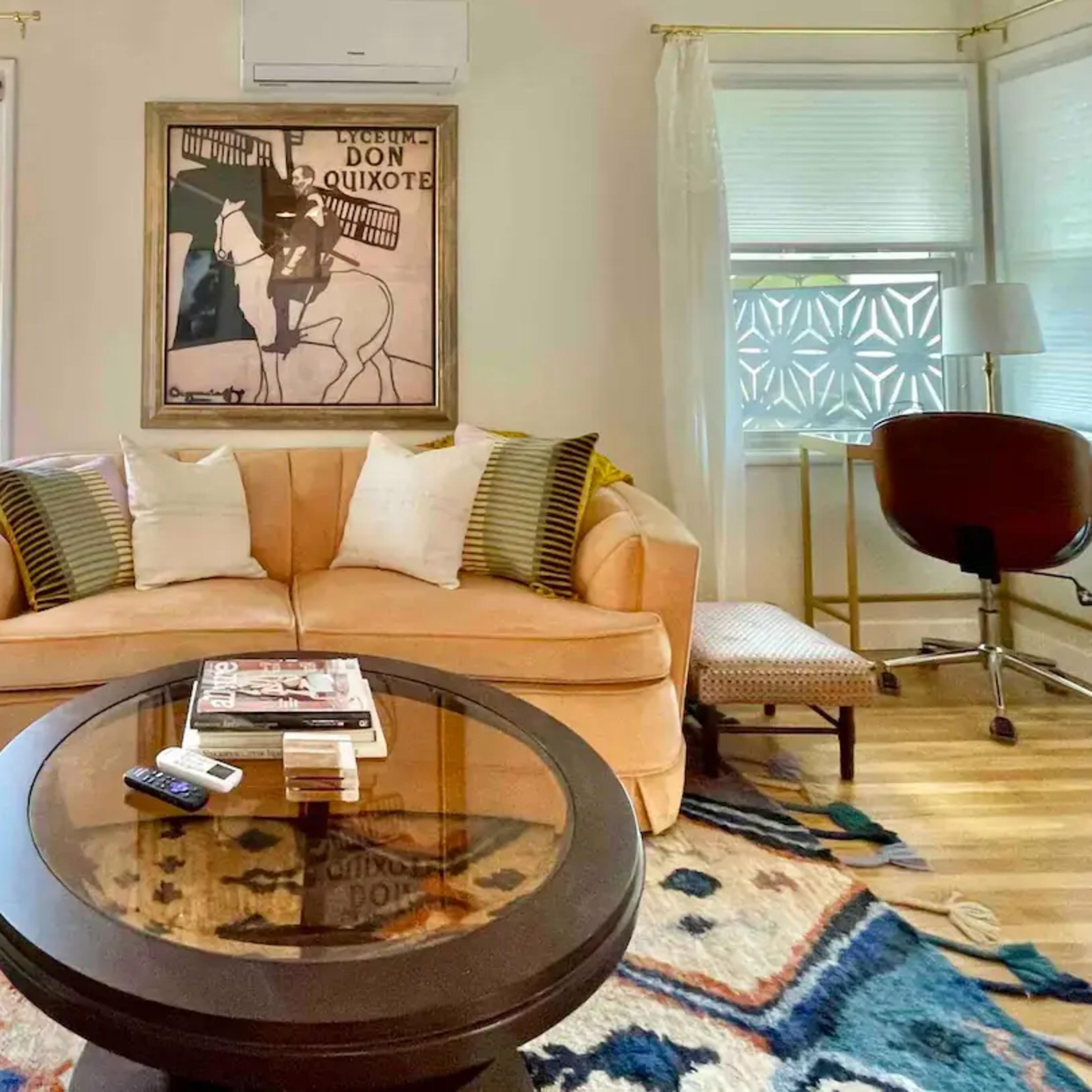 The living room features a peach-colored sofa, a round coffee table with magazines, and a framed artwork above, illuminated by natural light from the windows.