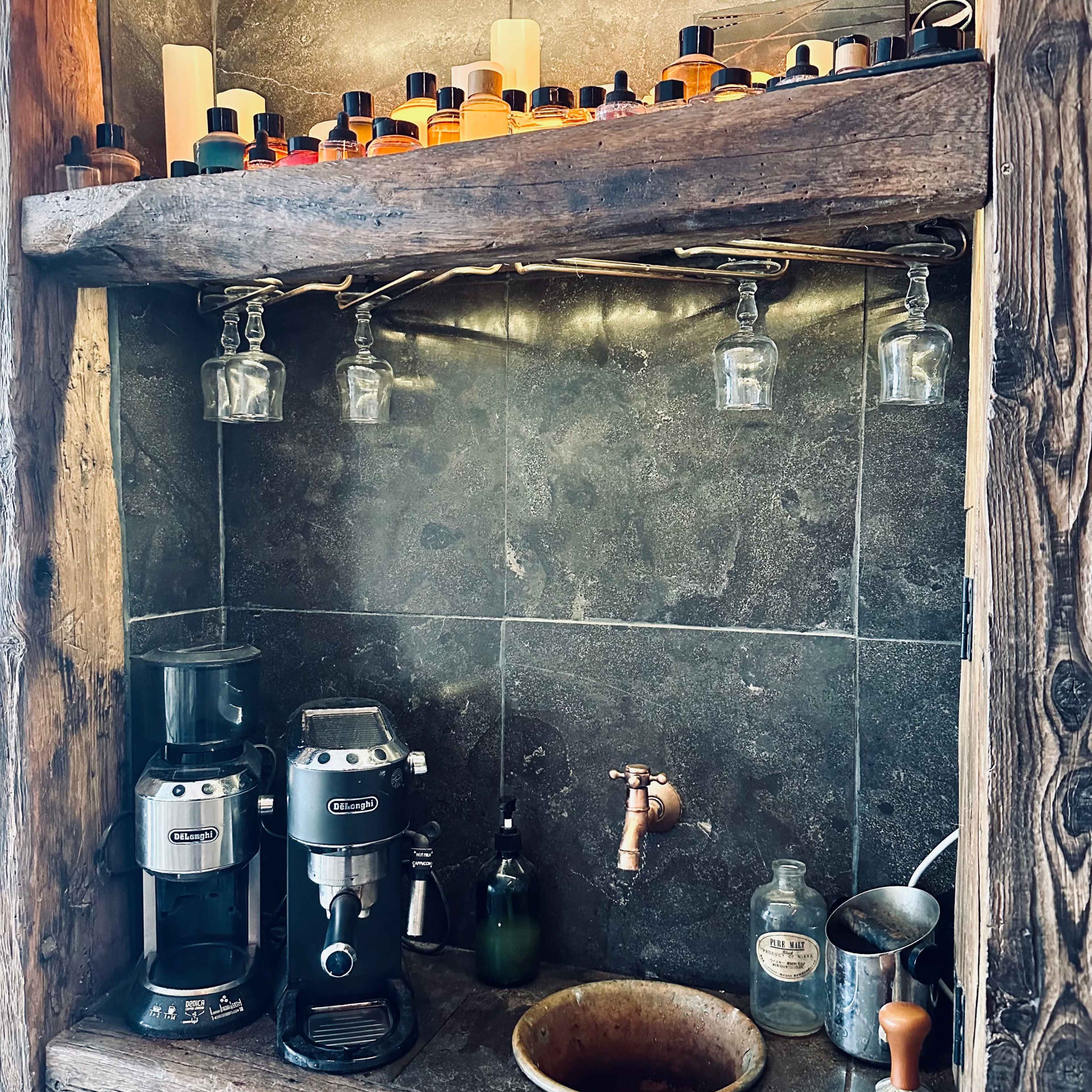 A rustic shelf with glass jars, a coffee machine, a sink with a faucet, and a bowl, set against a stone backdrop.