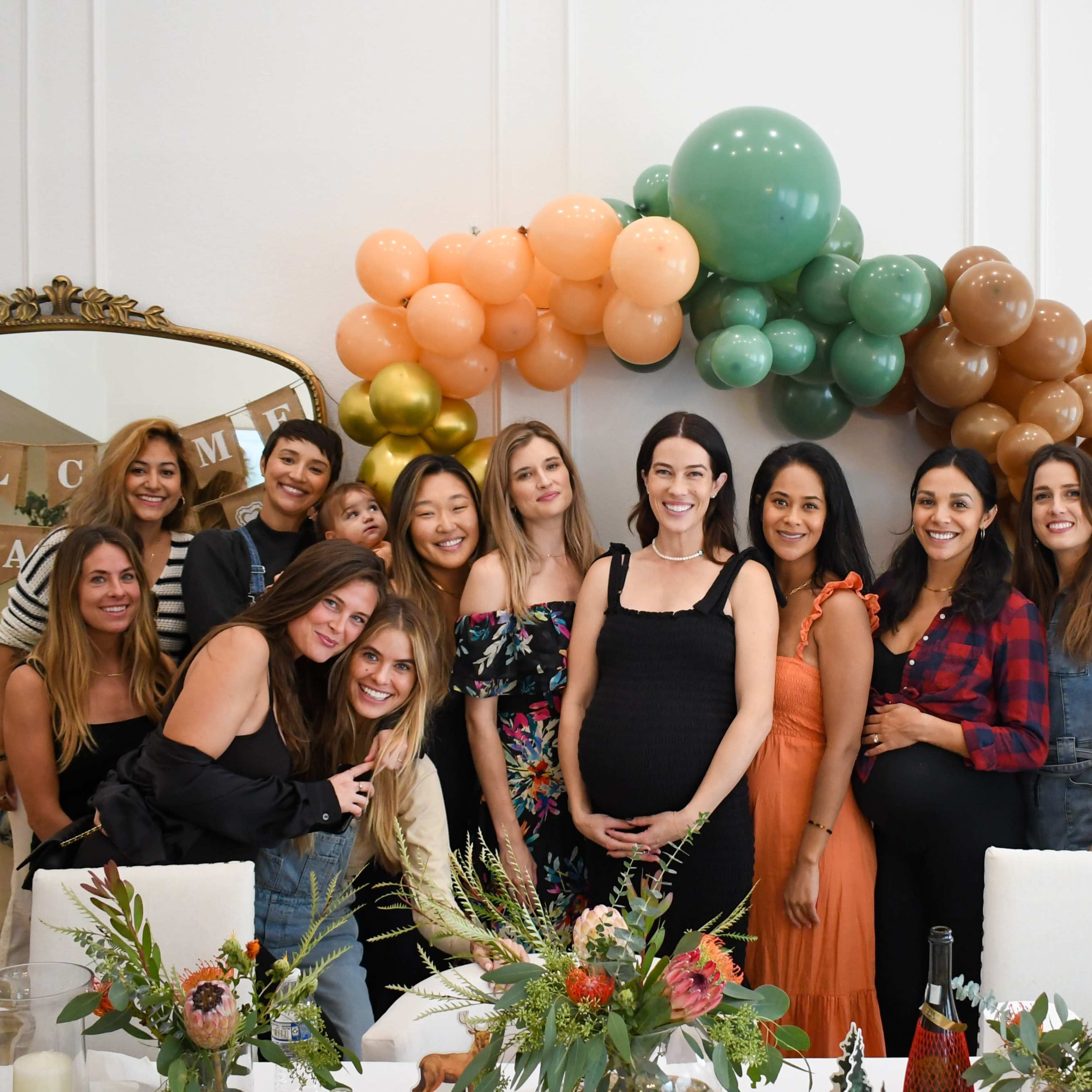 A group of twelve women stands together in a decorated room featuring a balloon arch, celebrating an event.