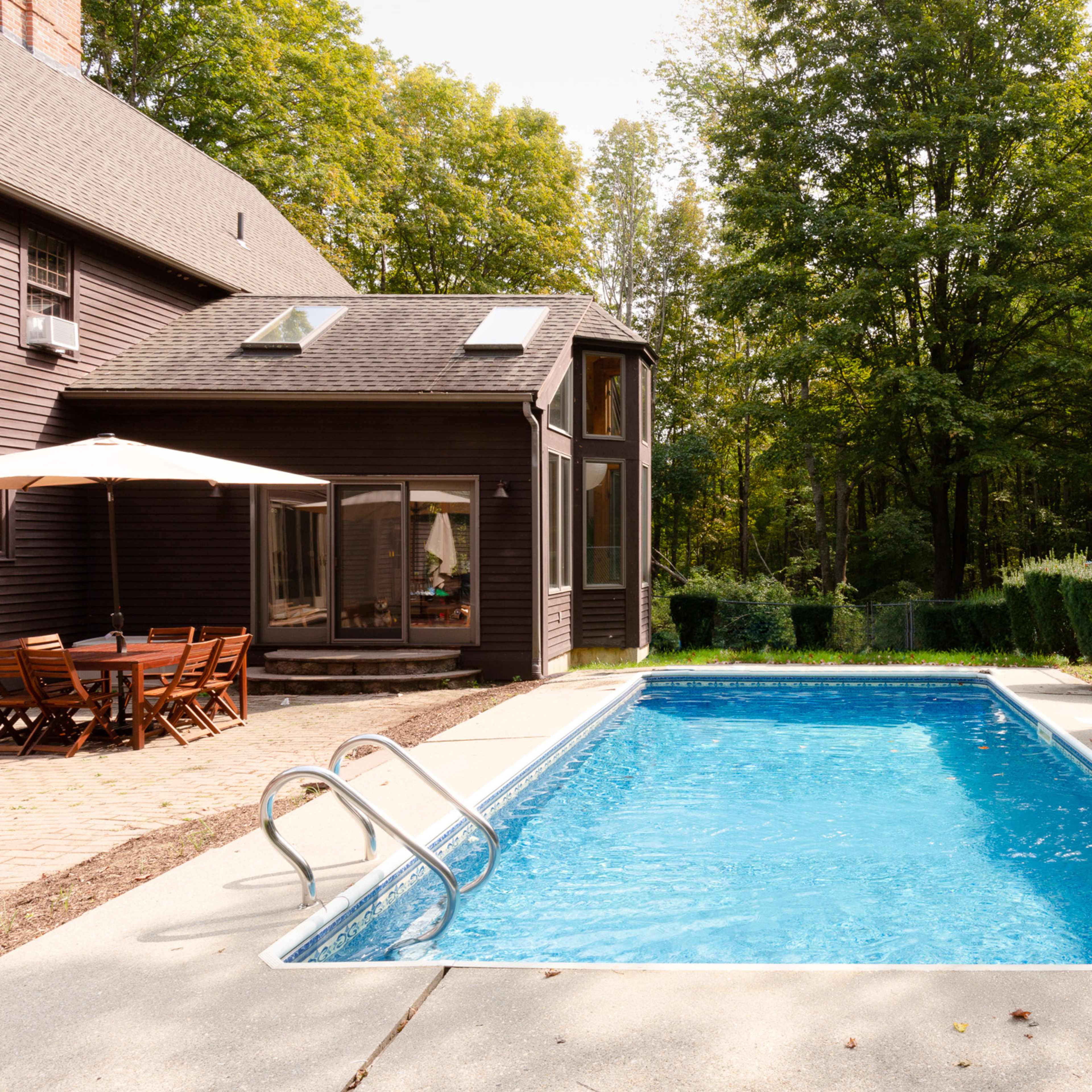A rectangular blue swimming pool surrounded by a concrete deck in a wooded backyard setting.
