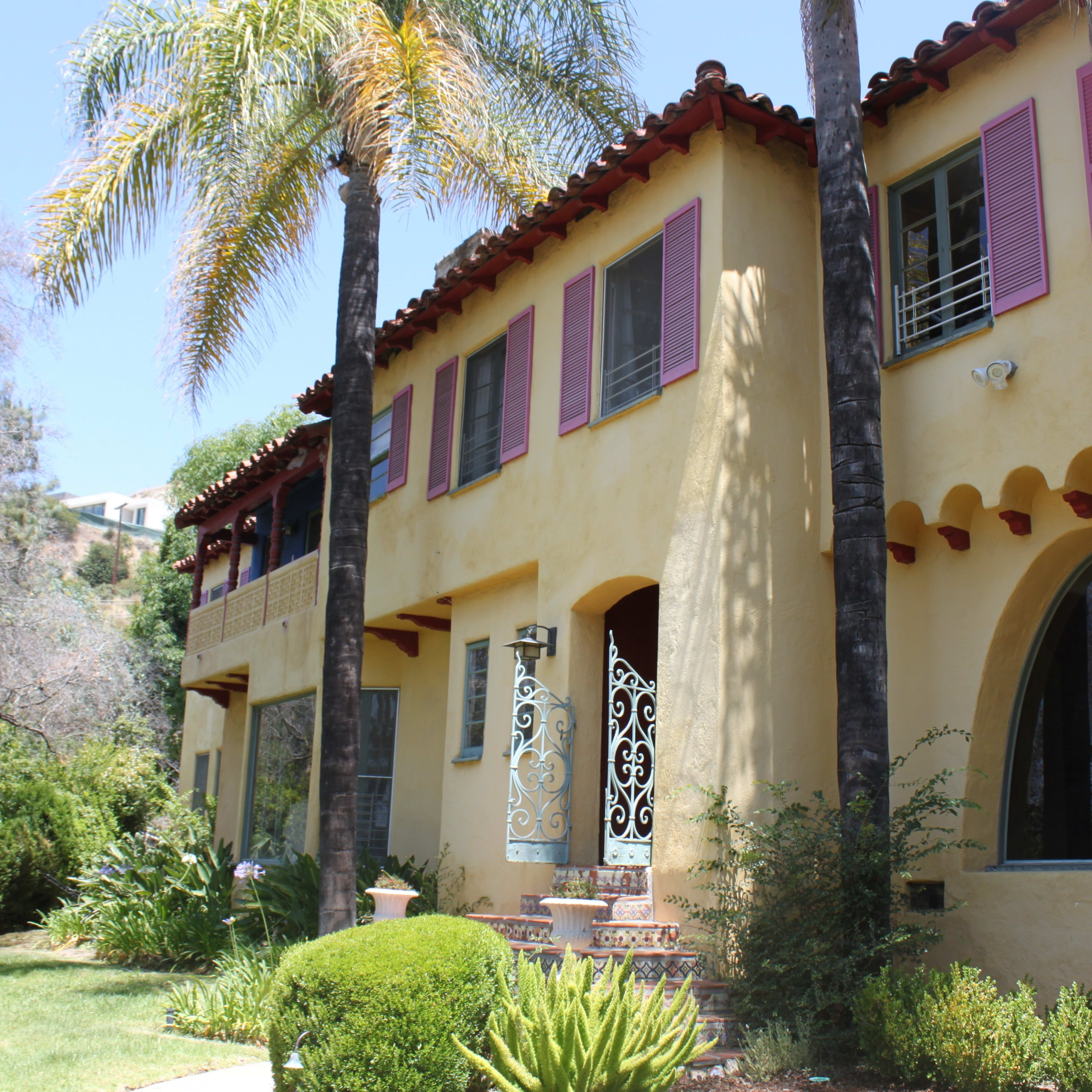 A two-story, yellow stucco house with decorative features, surrounded by lush greenery and palm trees.