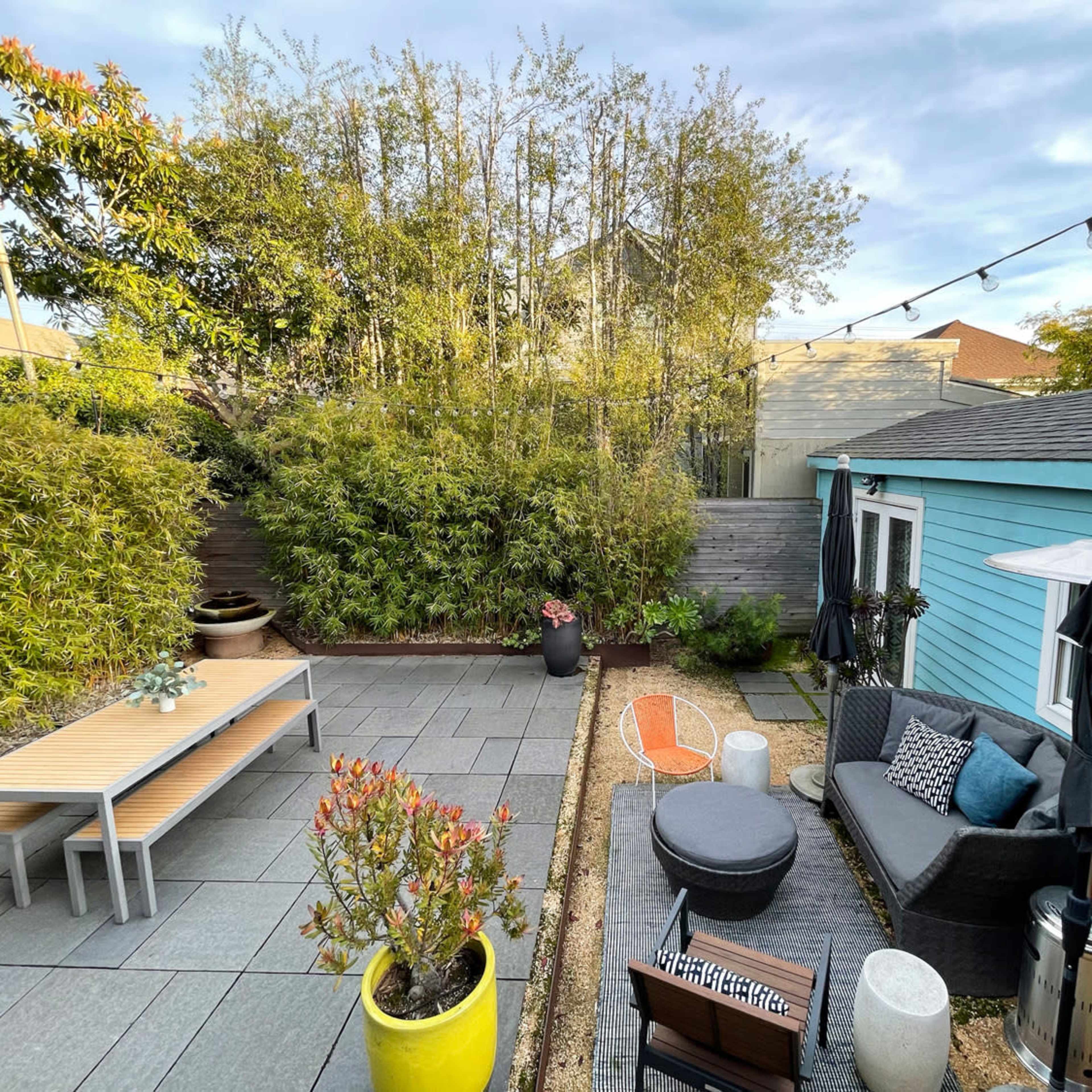 A landscaped backyard featuring a patio with a table, seating area, potted plants, and a blue shed in the background.