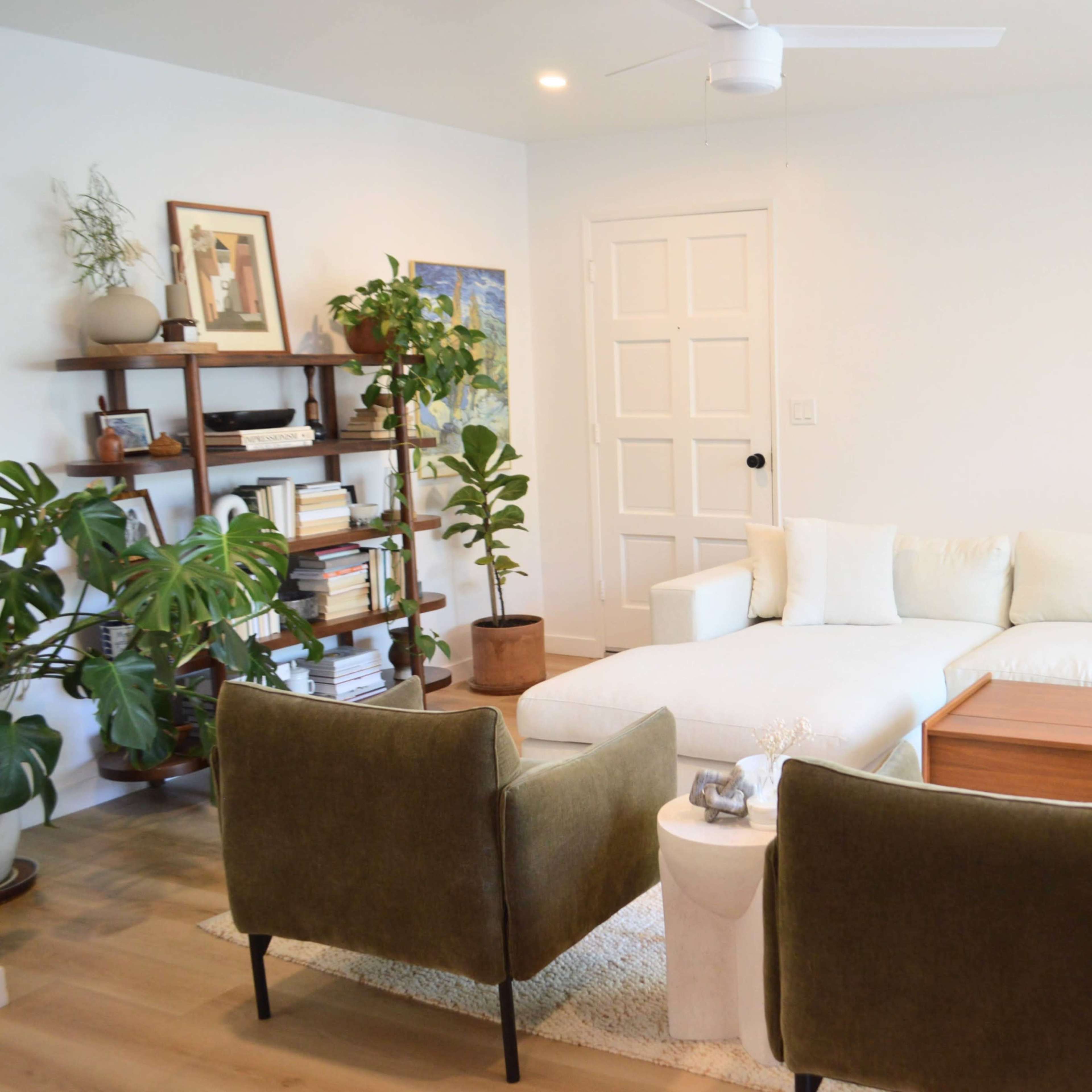 A brightly lit living room featuring a white sectional sofa, two green chairs, a wooden coffee table, and various plants on shelves and the floor.