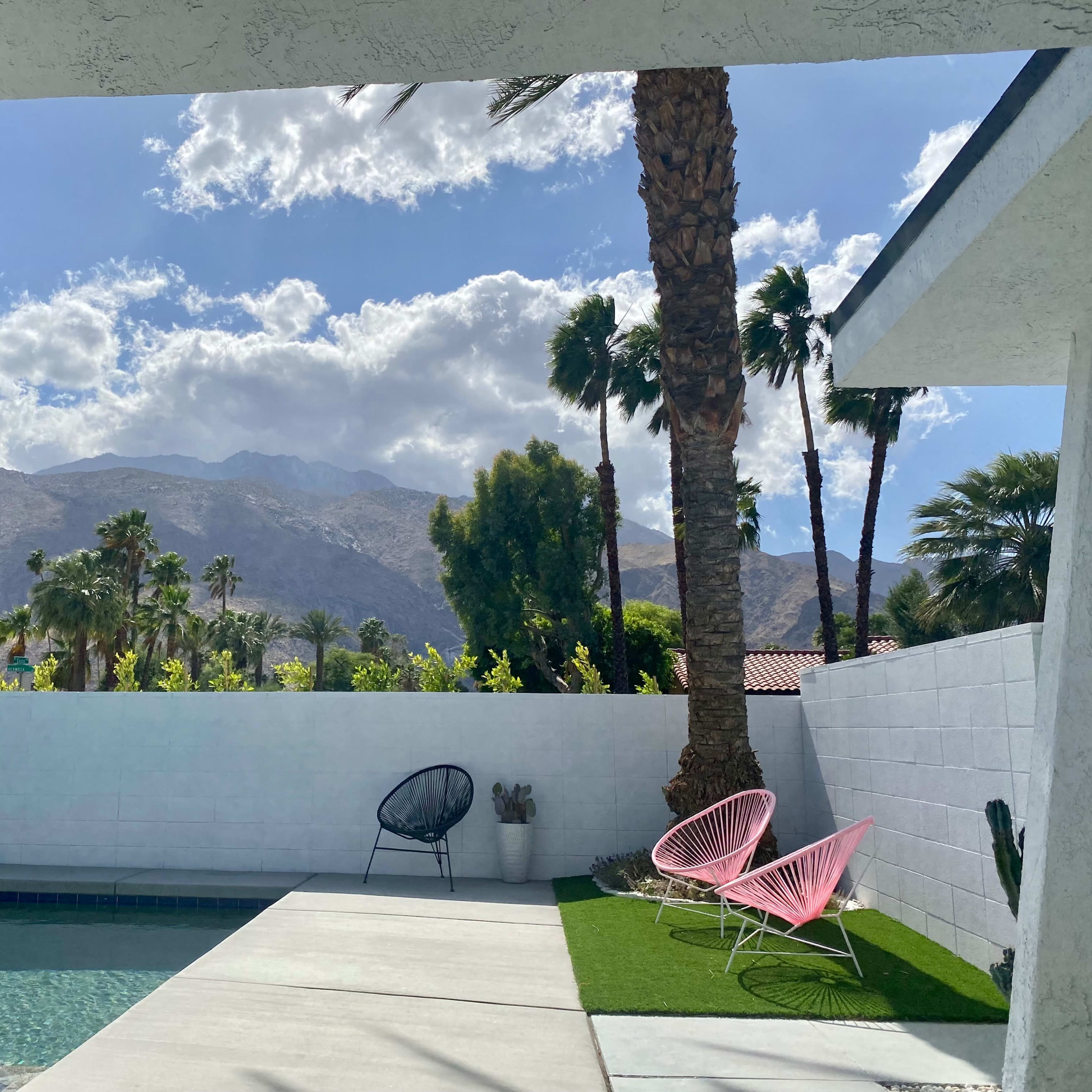 A poolside view with colorful chairs set against a backdrop of palm trees and mountains under a partly cloudy sky.