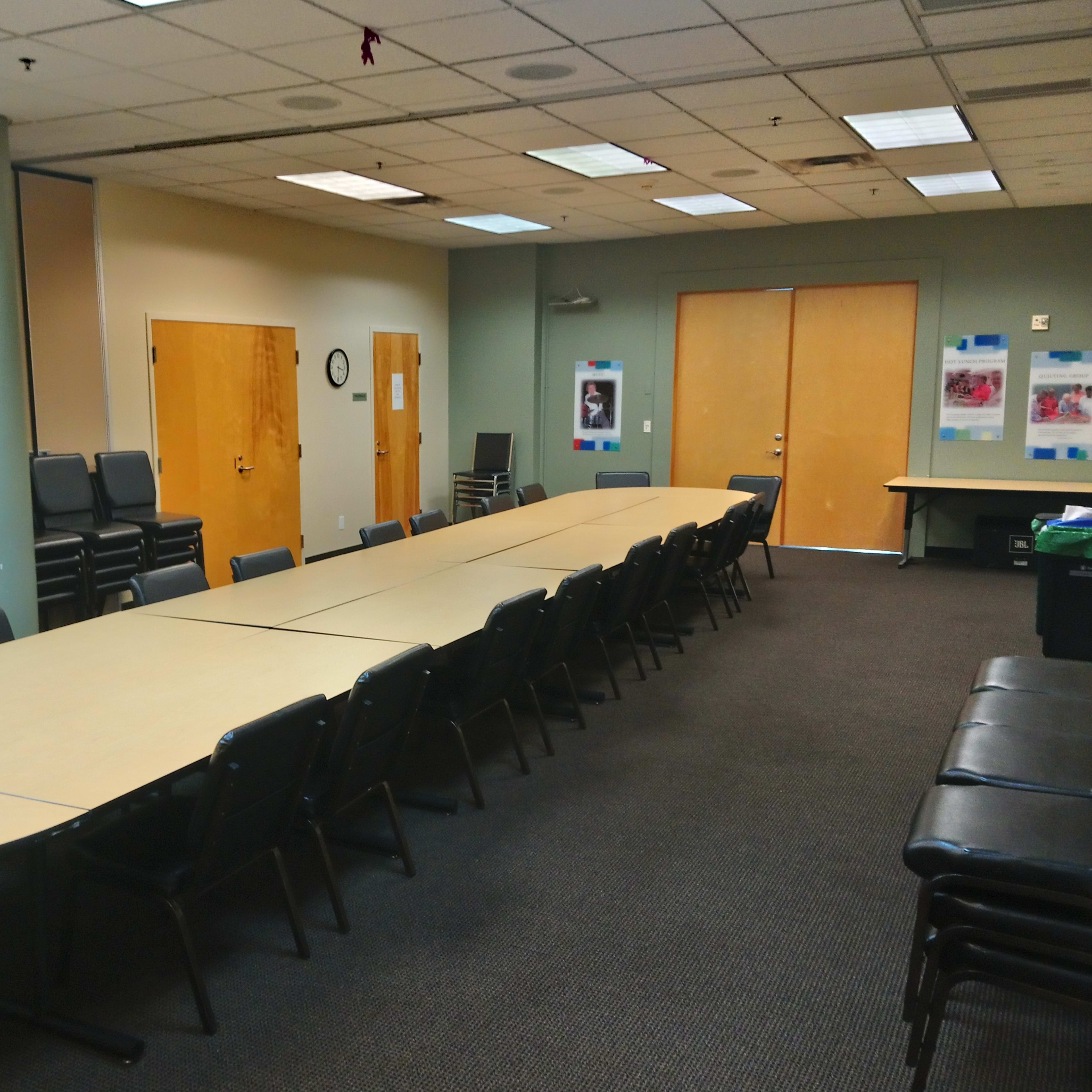 A long, oval conference table is set up in a meeting room with chairs lined along one side and a wall displaying posters.