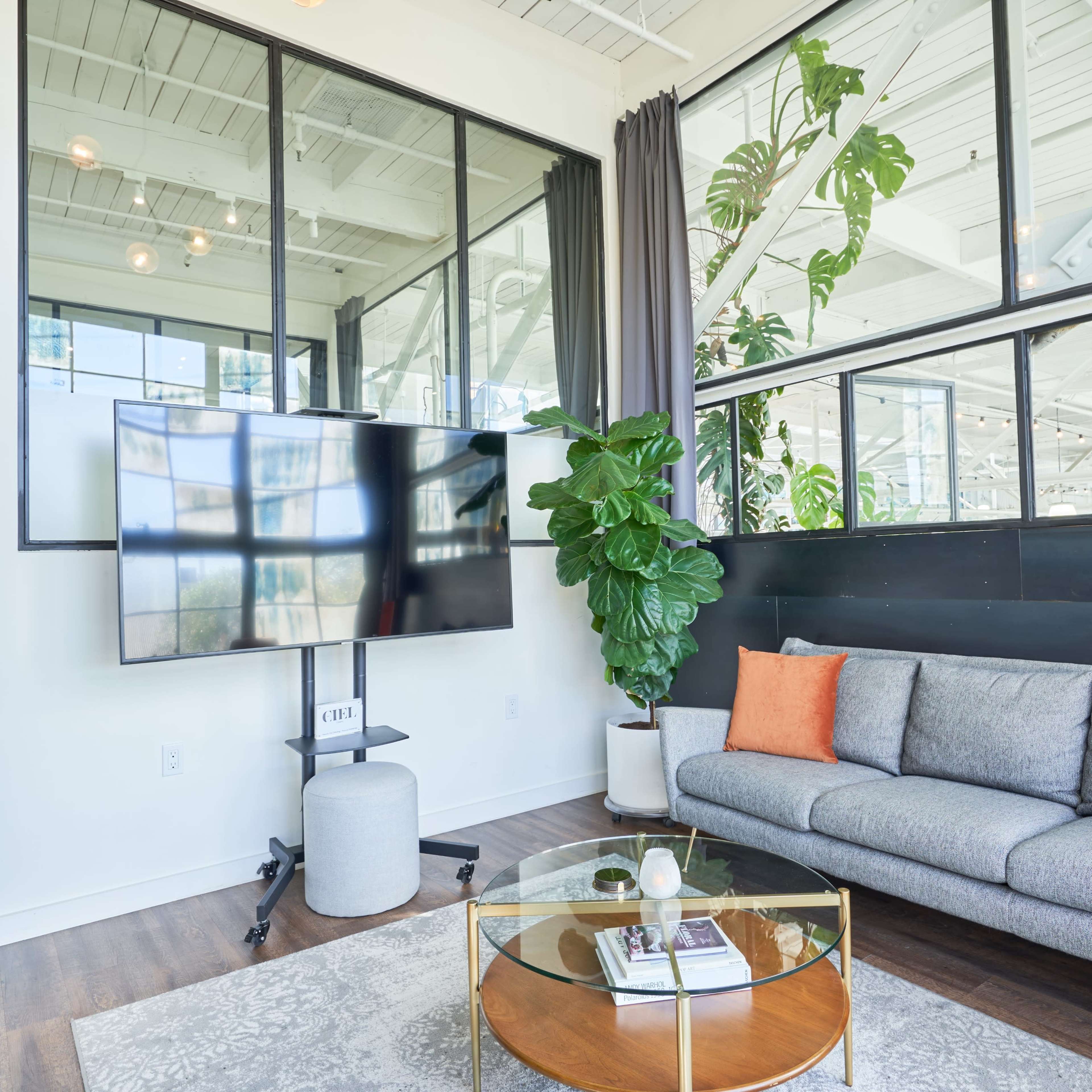 A modern living room with a gray sofa, a round coffee table, a large television mounted on the wall, and large windows that allow natural light to fill the space.