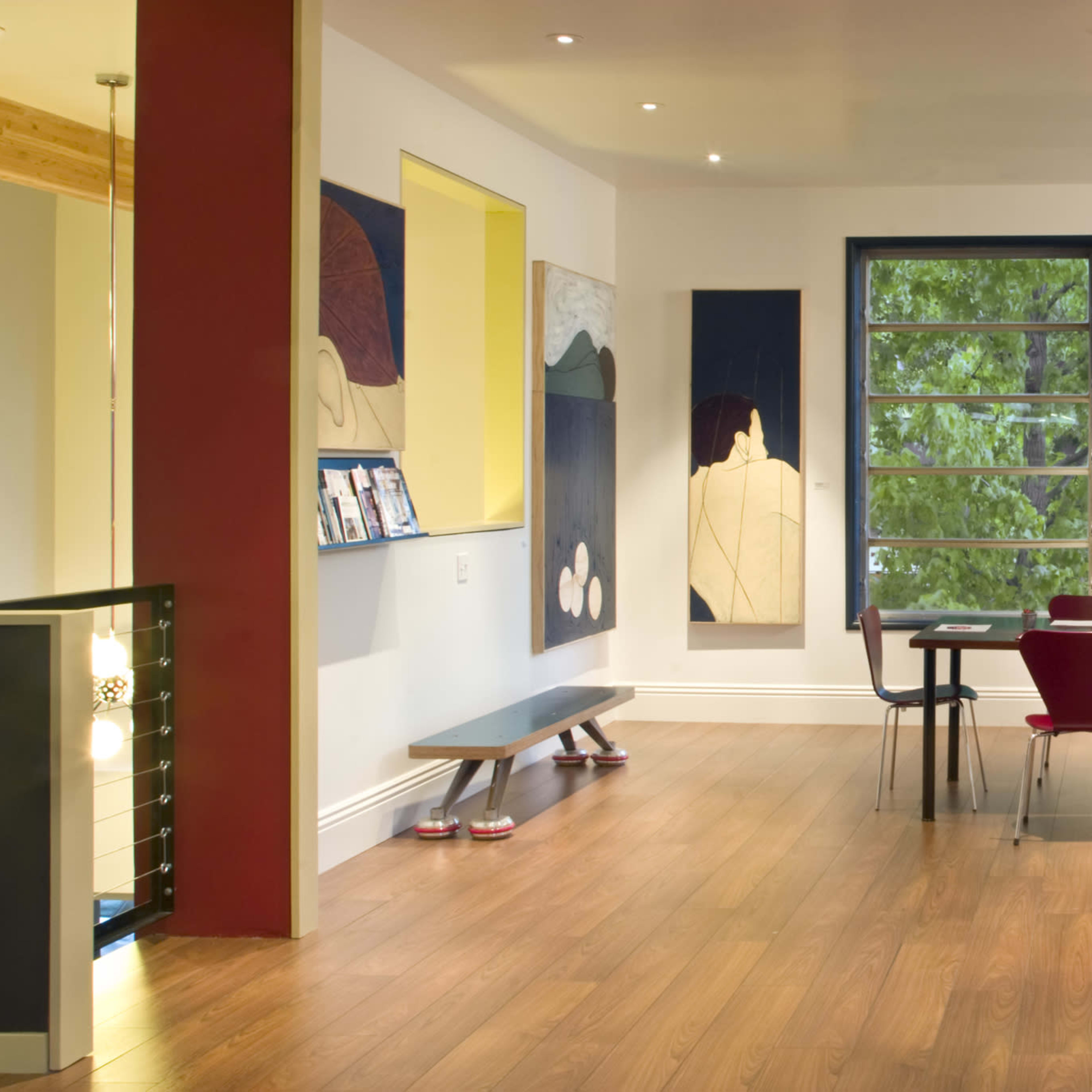 A modern interior space featuring artwork on the walls, wooden flooring, and a dining area with a table and red chairs near a large window.