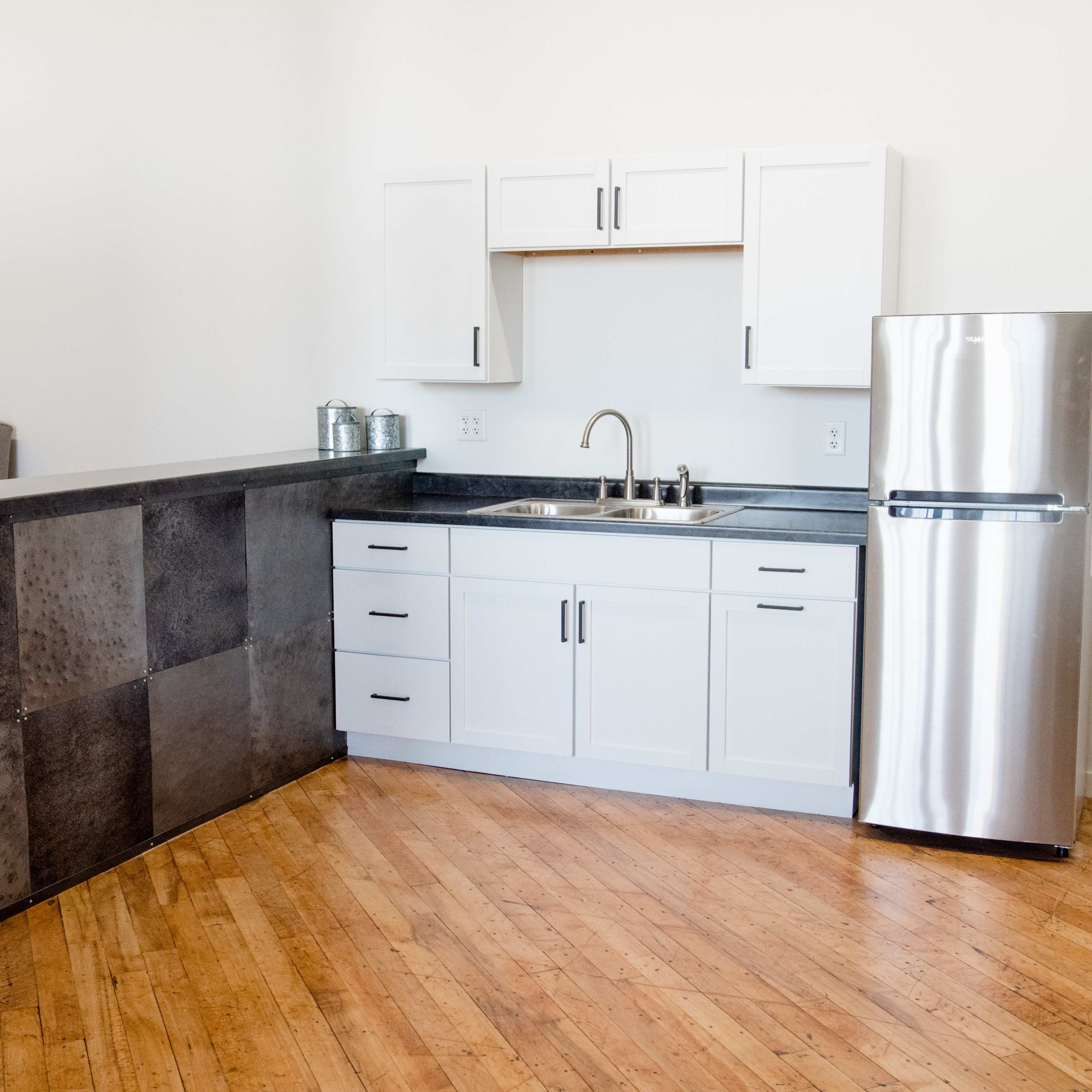 A modern kitchen area featuring a stainless steel refrigerator, white cabinets, a sink, and a dark countertop, with wooden flooring.