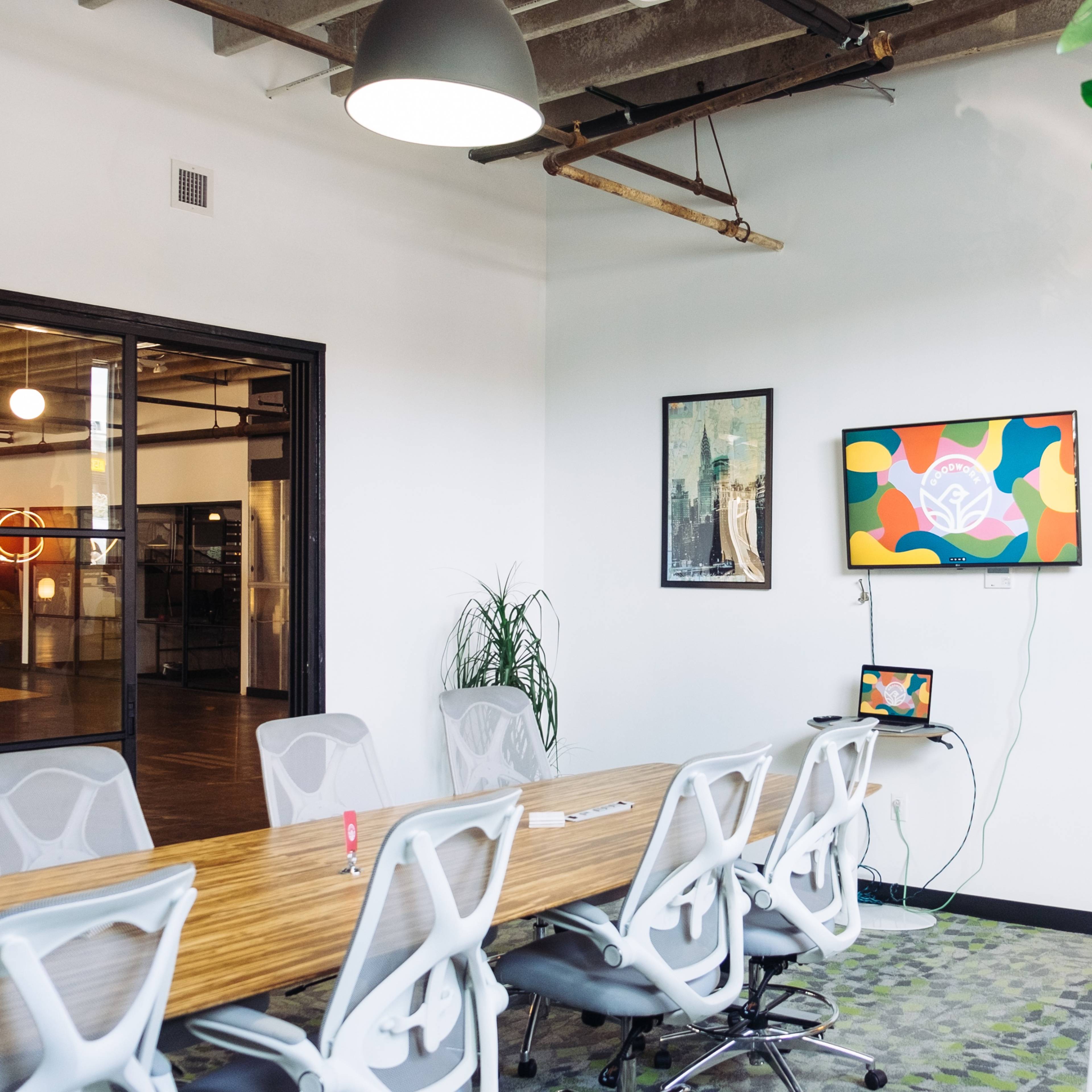 A modern conference room features a long wooden table surrounded by ergonomic chairs, with a colorful artwork displayed on a wall-mounted screen.