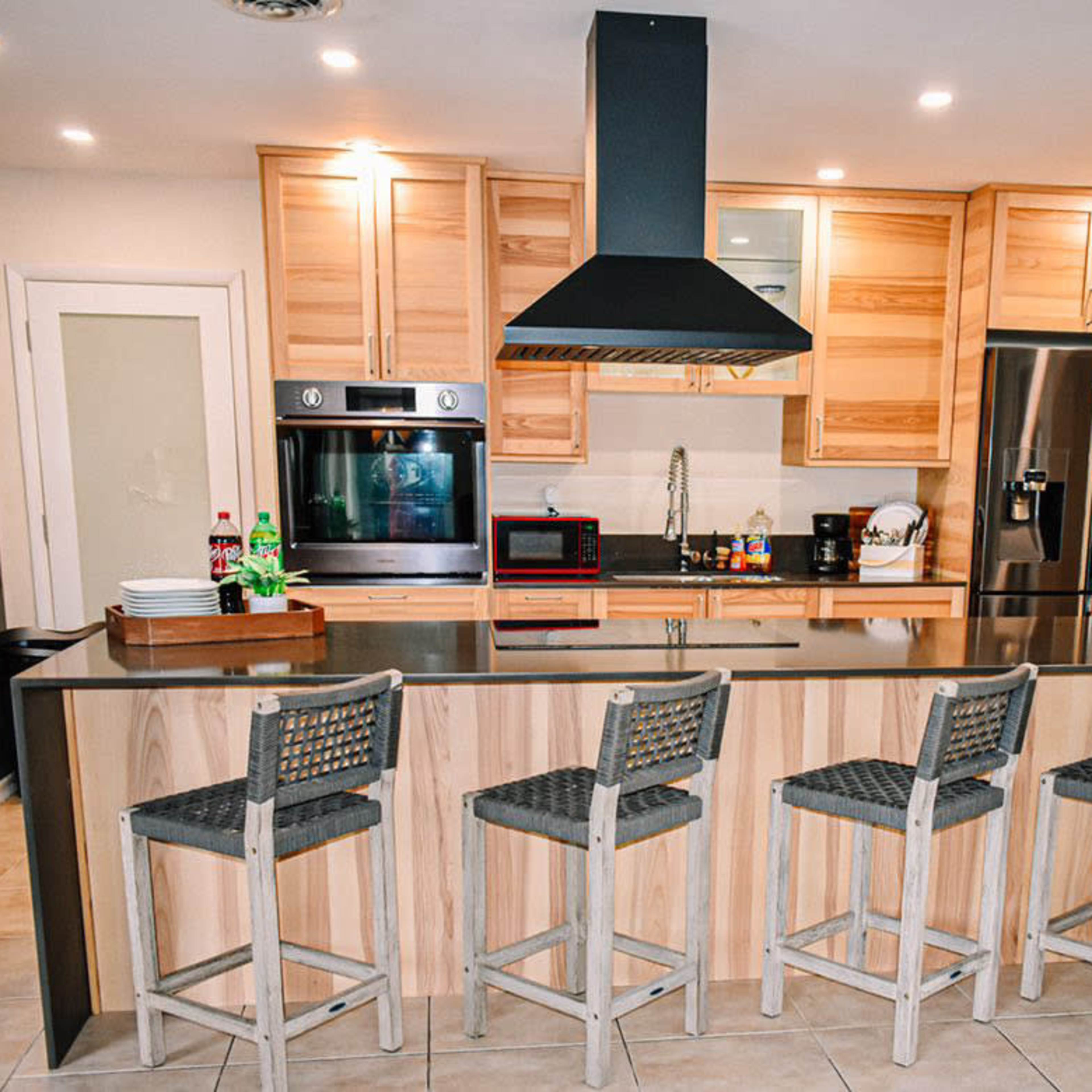 A modern kitchen featuring a large island with four barstools, wooden cabinetry, stainless steel appliances, and a range hood above the cooking area.