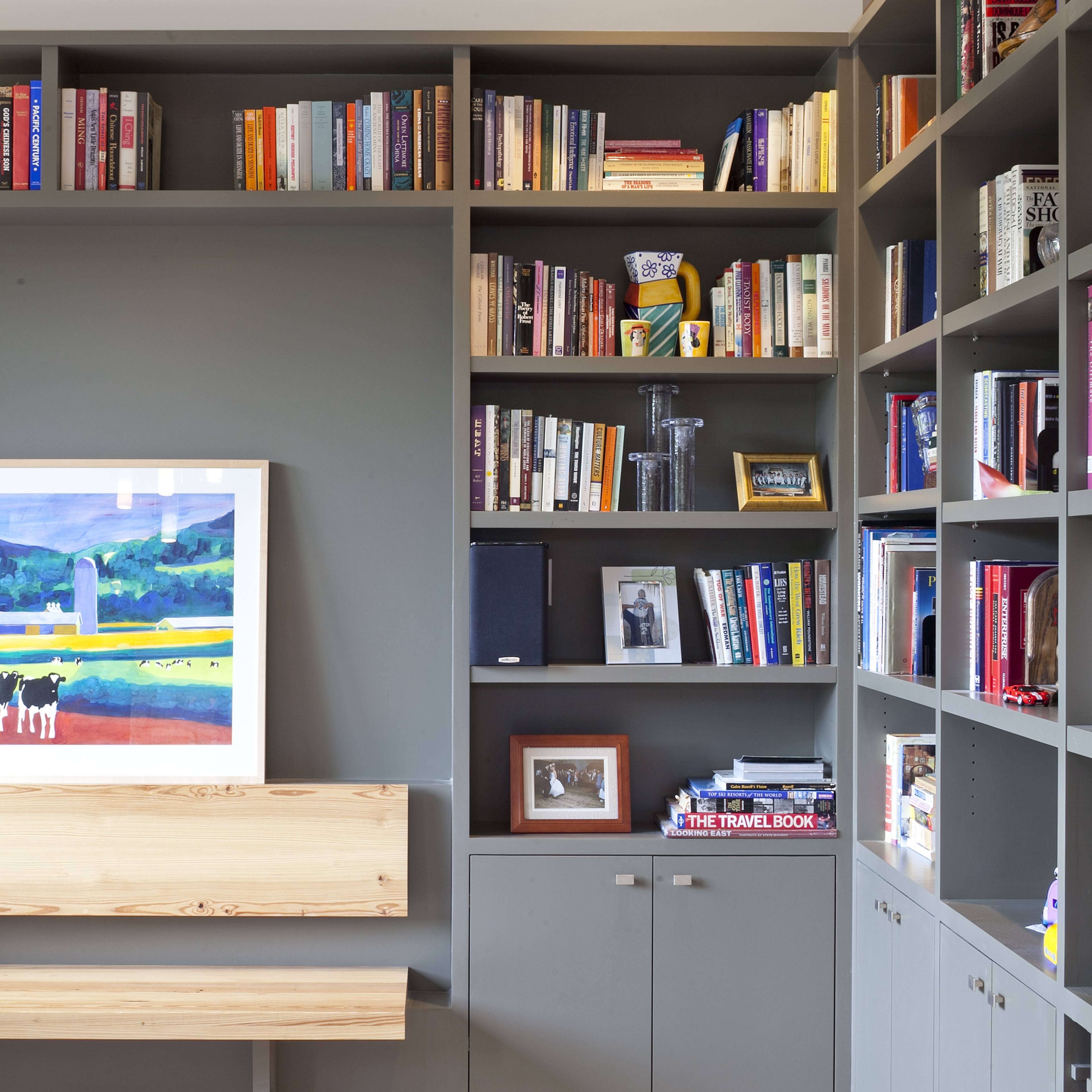 A modern bookshelf filled with books and framed pictures is complemented by a wooden bench against a gray wall.
