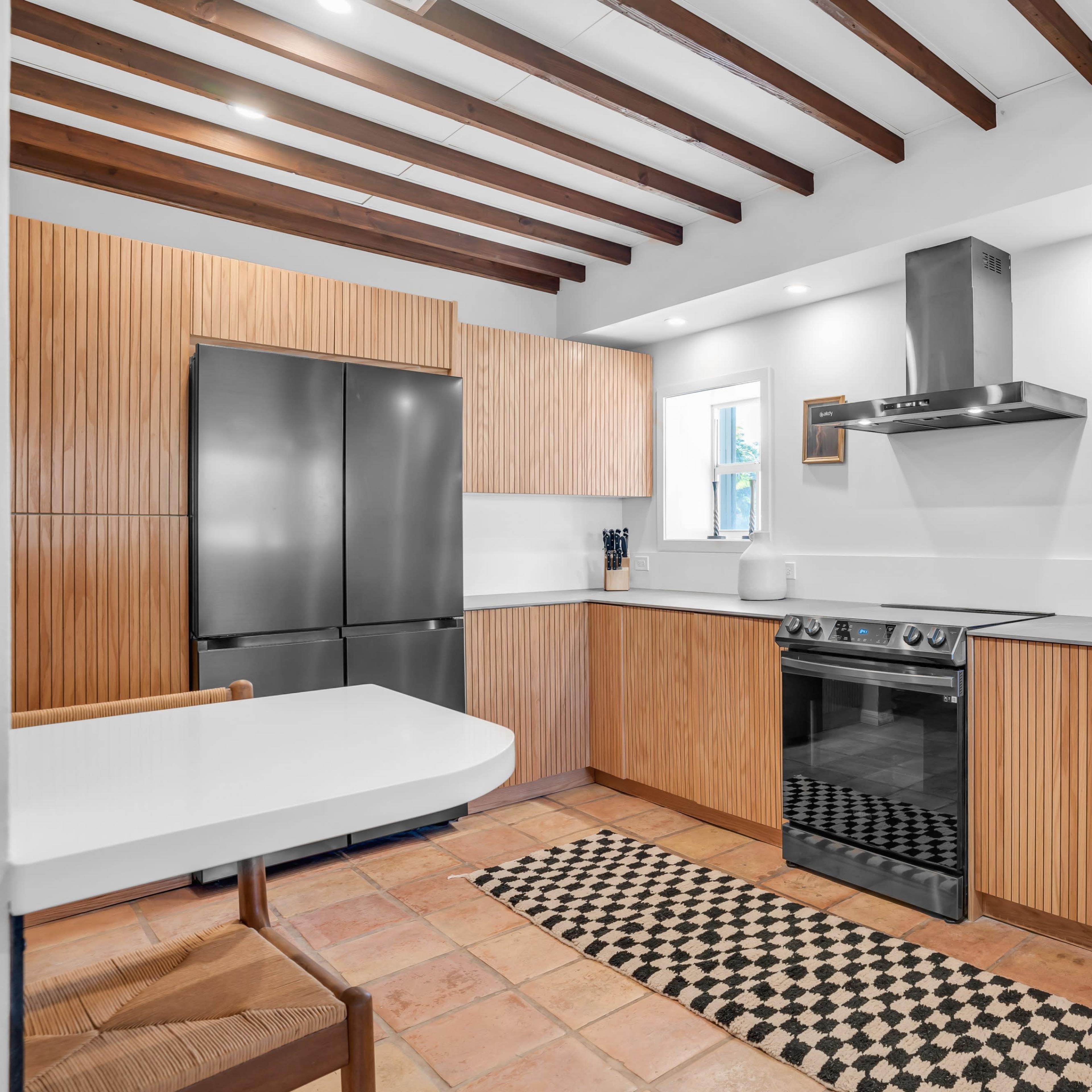 A modern kitchen features wooden cabinetry, stainless steel appliances, and a tiled floor with a checkered rug.