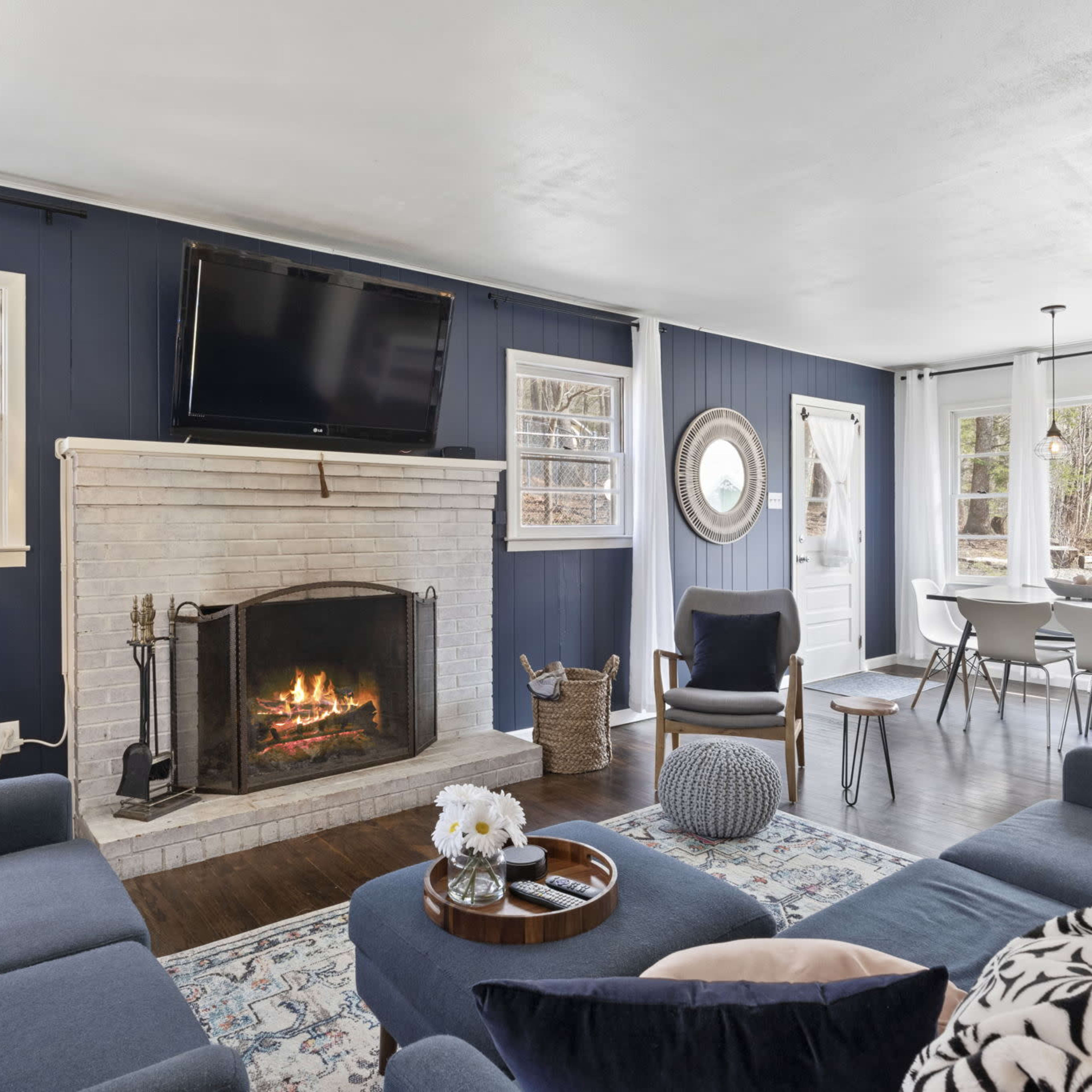 A cozy living room with a fireplace, a mounted TV, and a dining area adjacent to large windows overlooking a natural landscape.