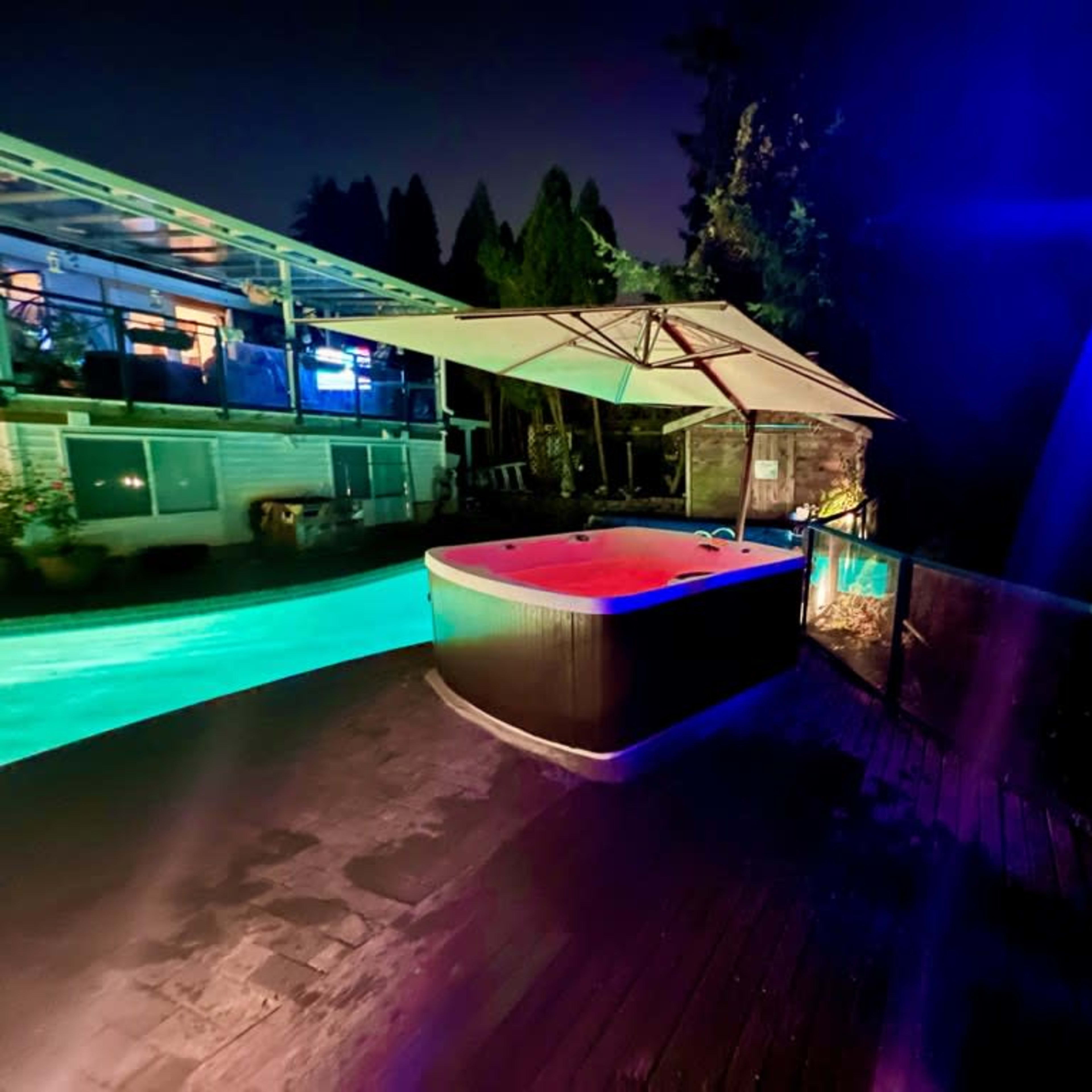 A hot tub with colorful lighting on a wooden deck next to a swimming pool, surrounded by trees at night.