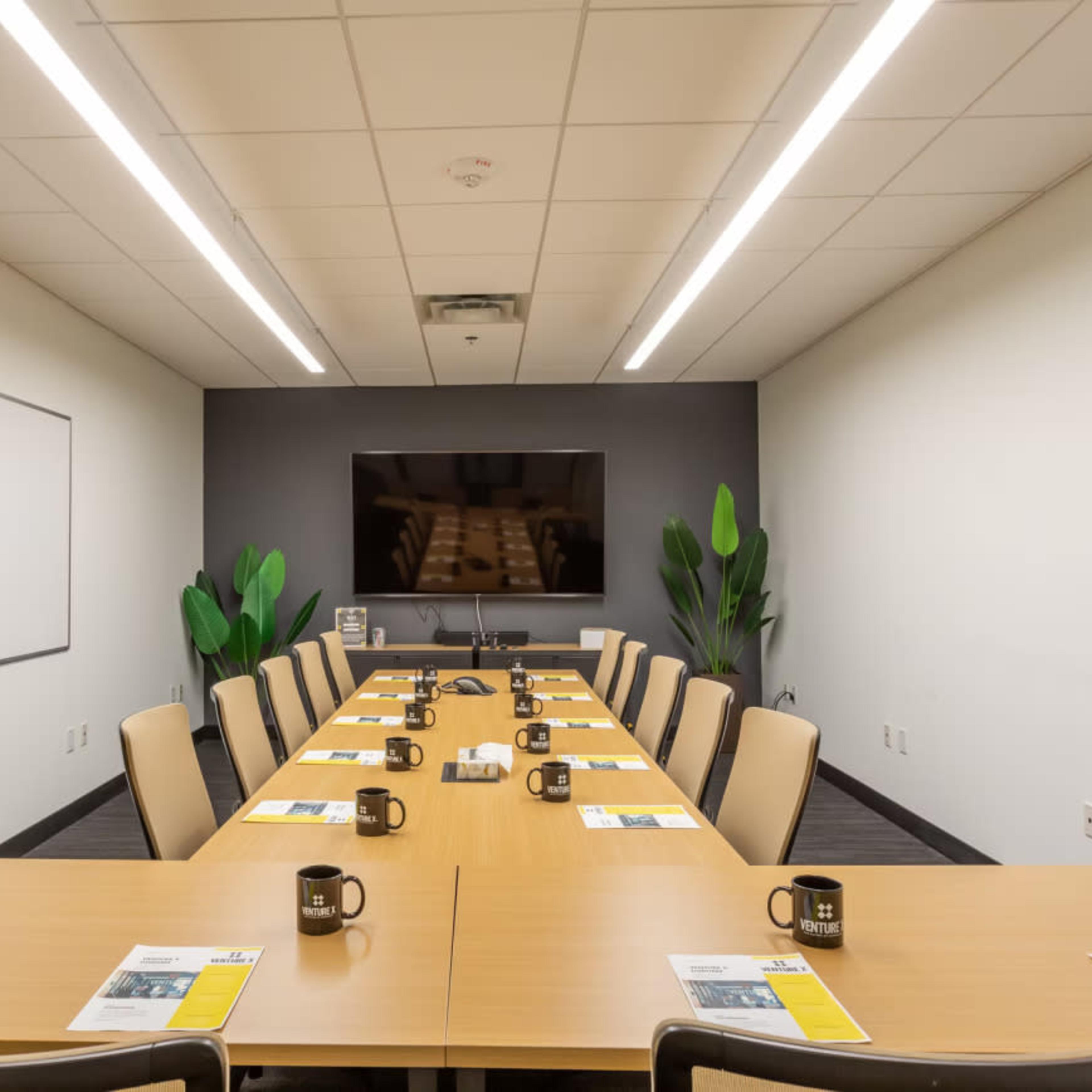 A conference room table is set up for a meeting, with place settings, mugs, and a large screen at one end.