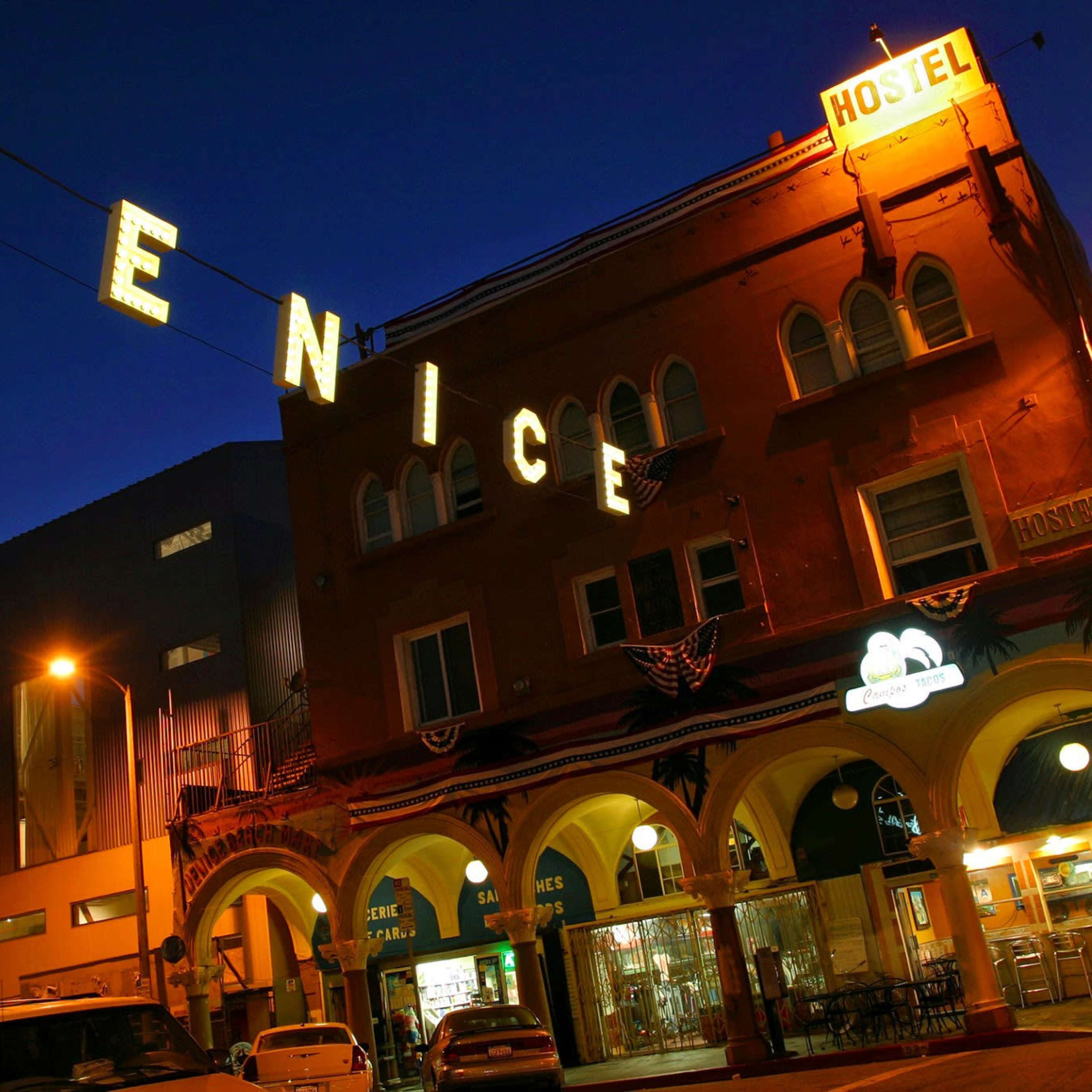 A brightly lit hostel with "VENICE" signage is seen at night, located in a building featuring arched entrances and decorative elements.