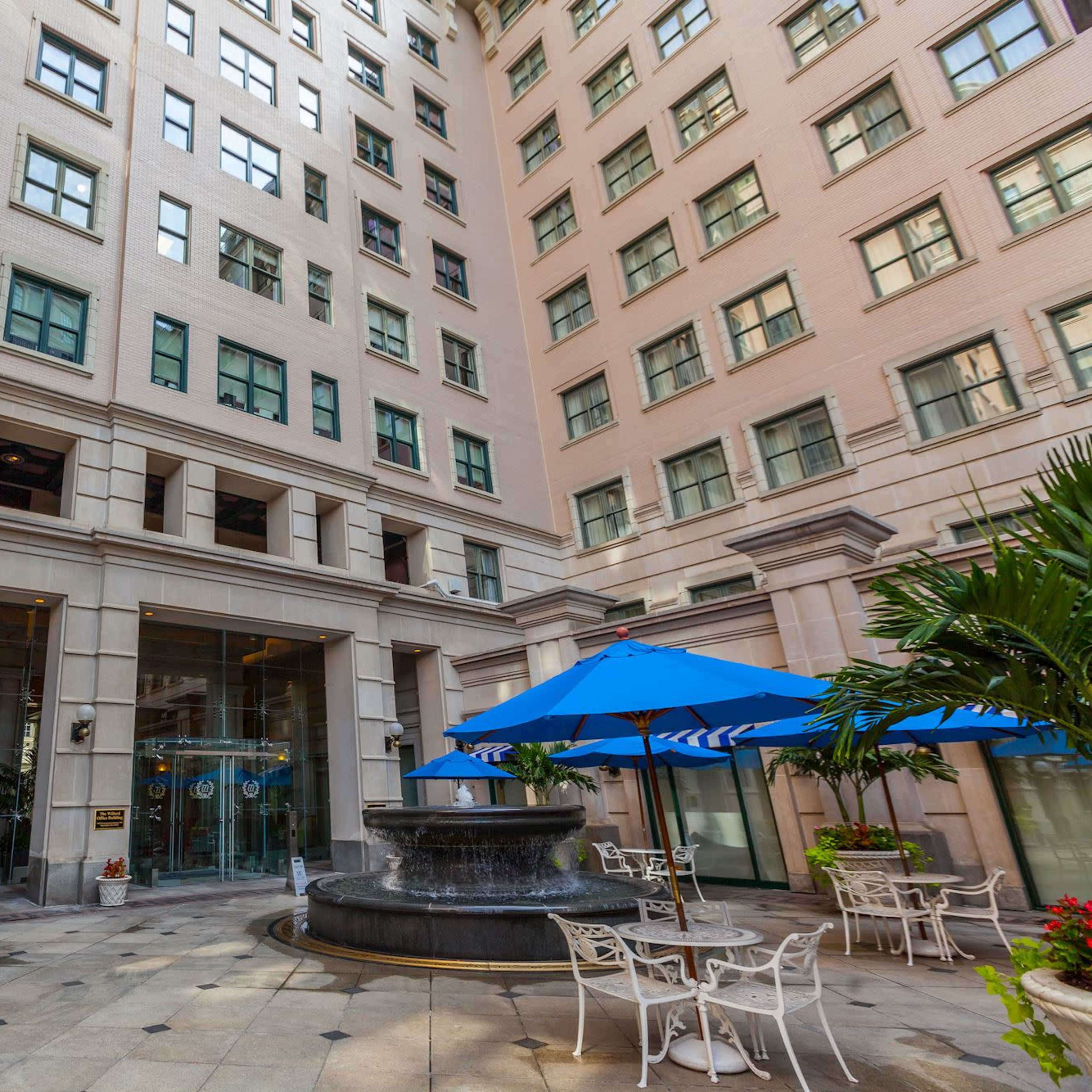 A courtyard featuring a fountain, surrounded by tables and chairs under blue umbrellas, with a tall building in the background.