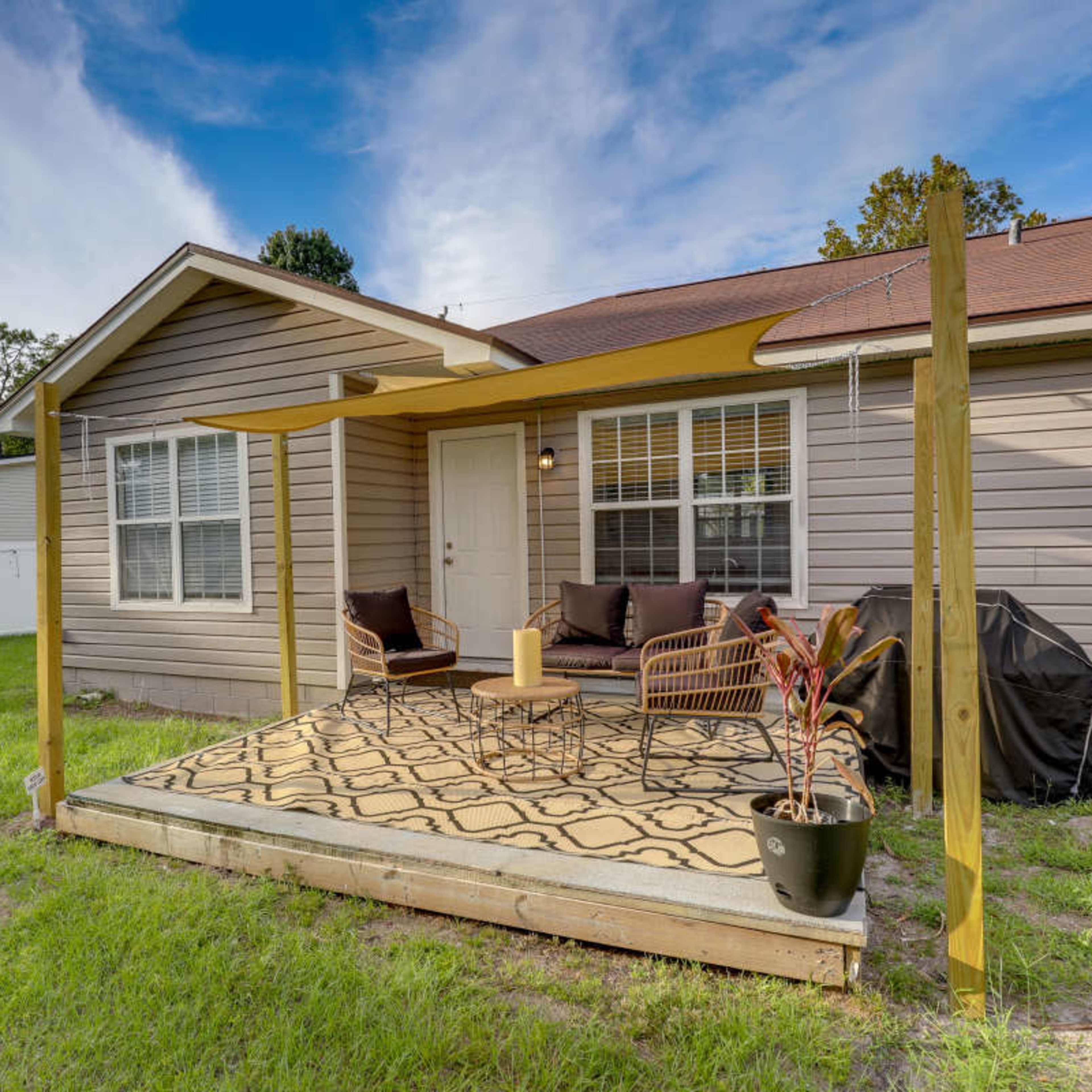 A small wooden porch with seating furniture, a decorative rug, and a potted plant, adjacent to a single-story house surrounded by grass.