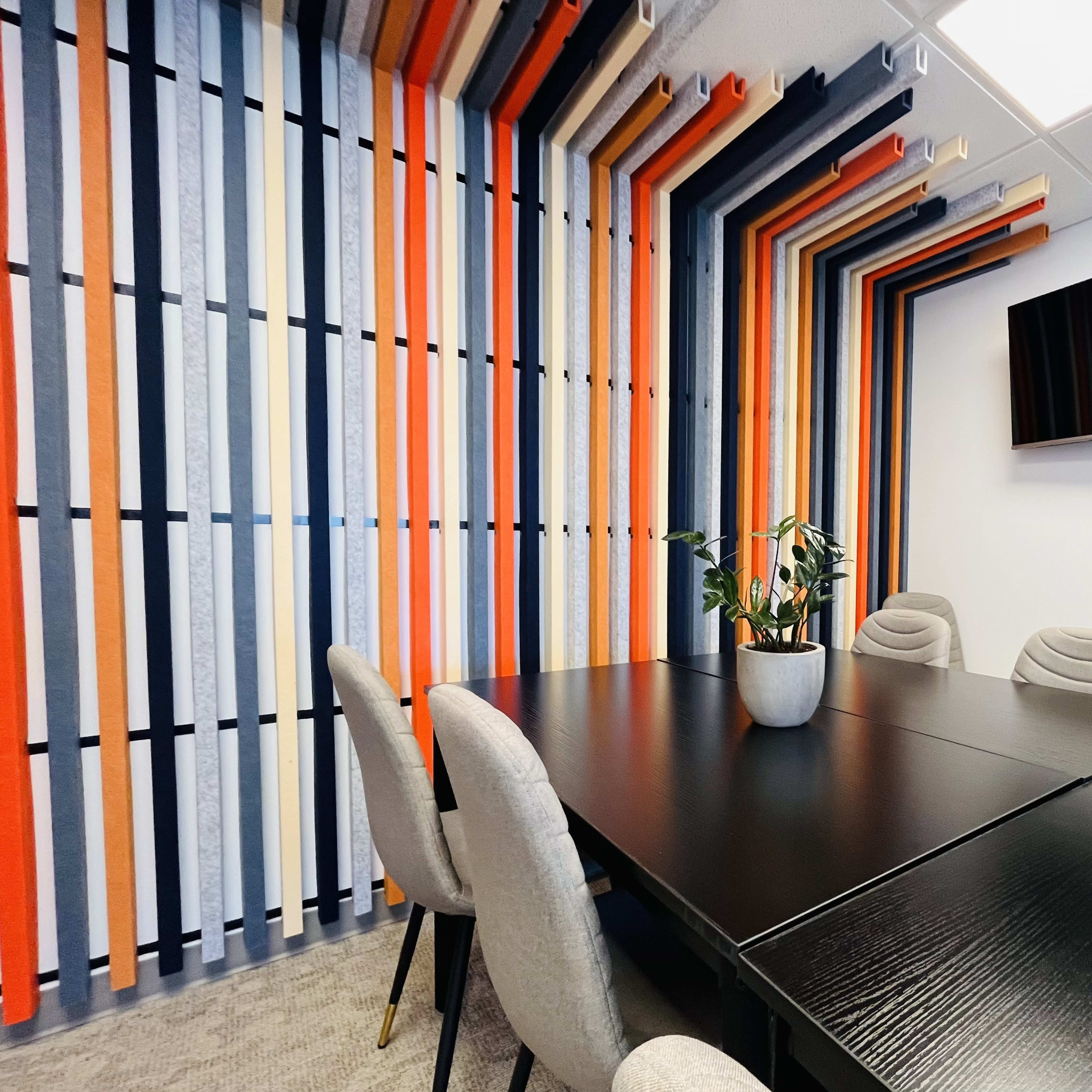 A conference room features a black table surrounded by gray chairs and a wall adorned with colorful vertical stripes, with a television mounted above.
