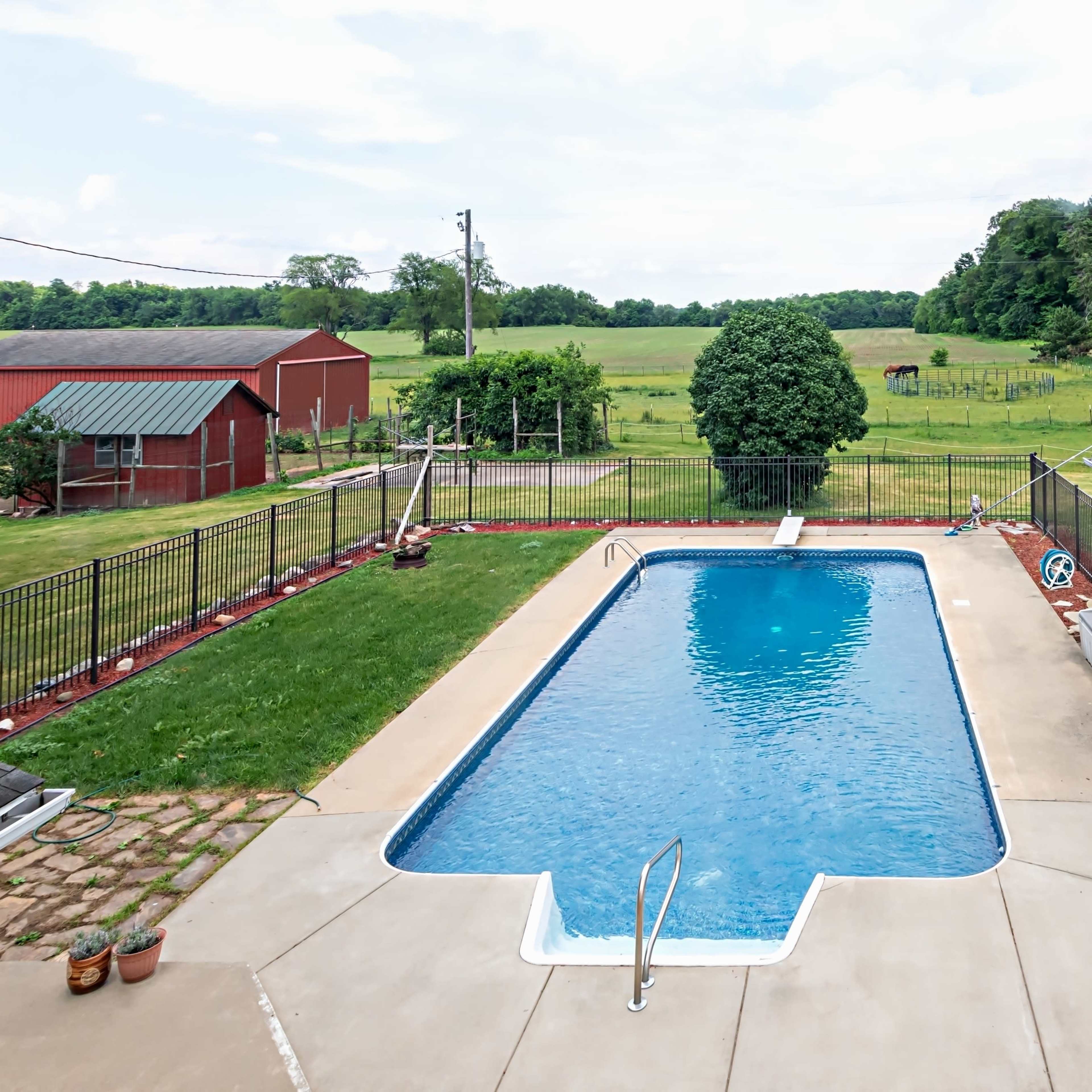 A rectangular swimming pool surrounded by a concrete deck, with a fenced yard and a red barn visible in the background.