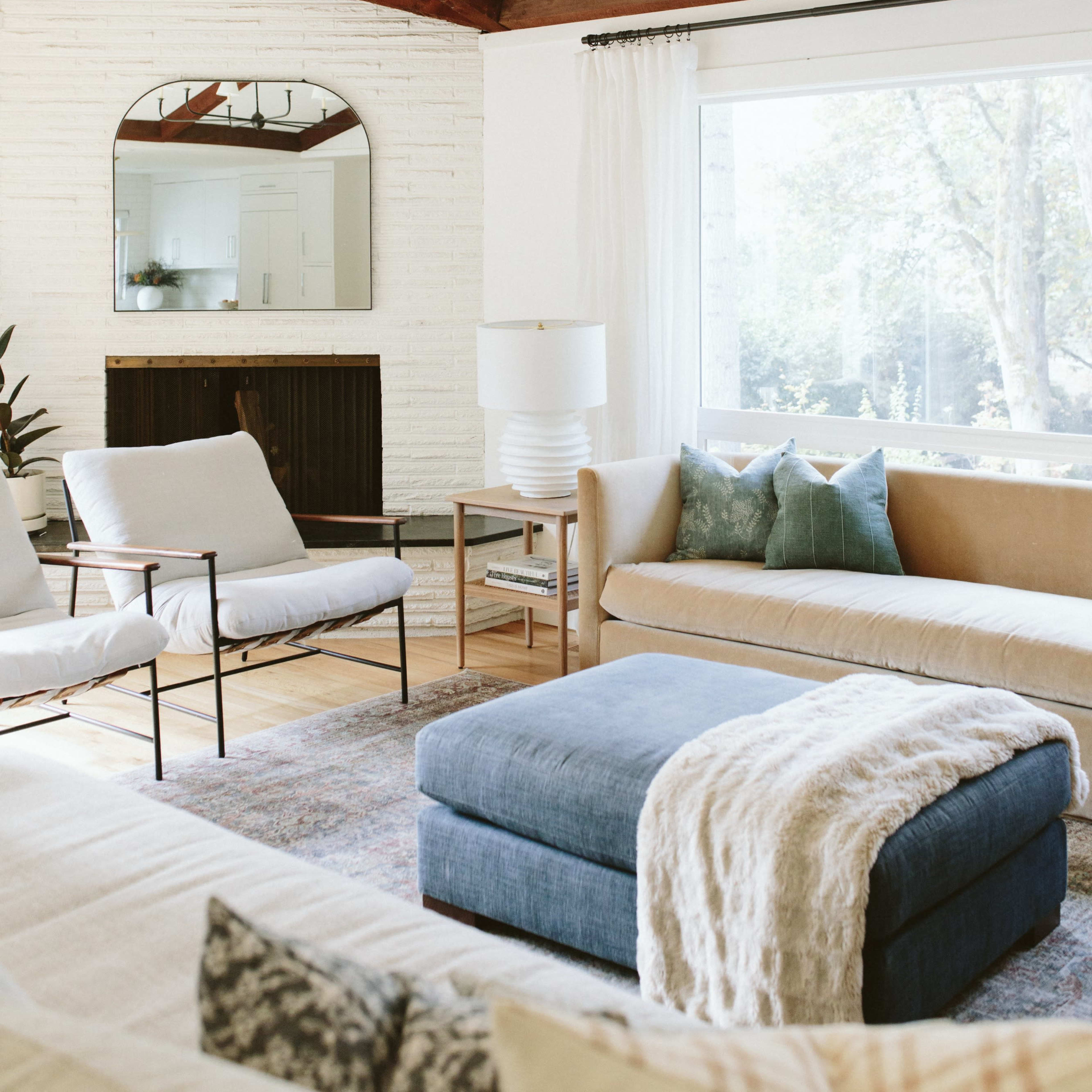The living room features a light-colored sofa, a blue ottoman, and two armchairs, with large windows allowing natural light to fill the space, complemented by wooden beams overhead.