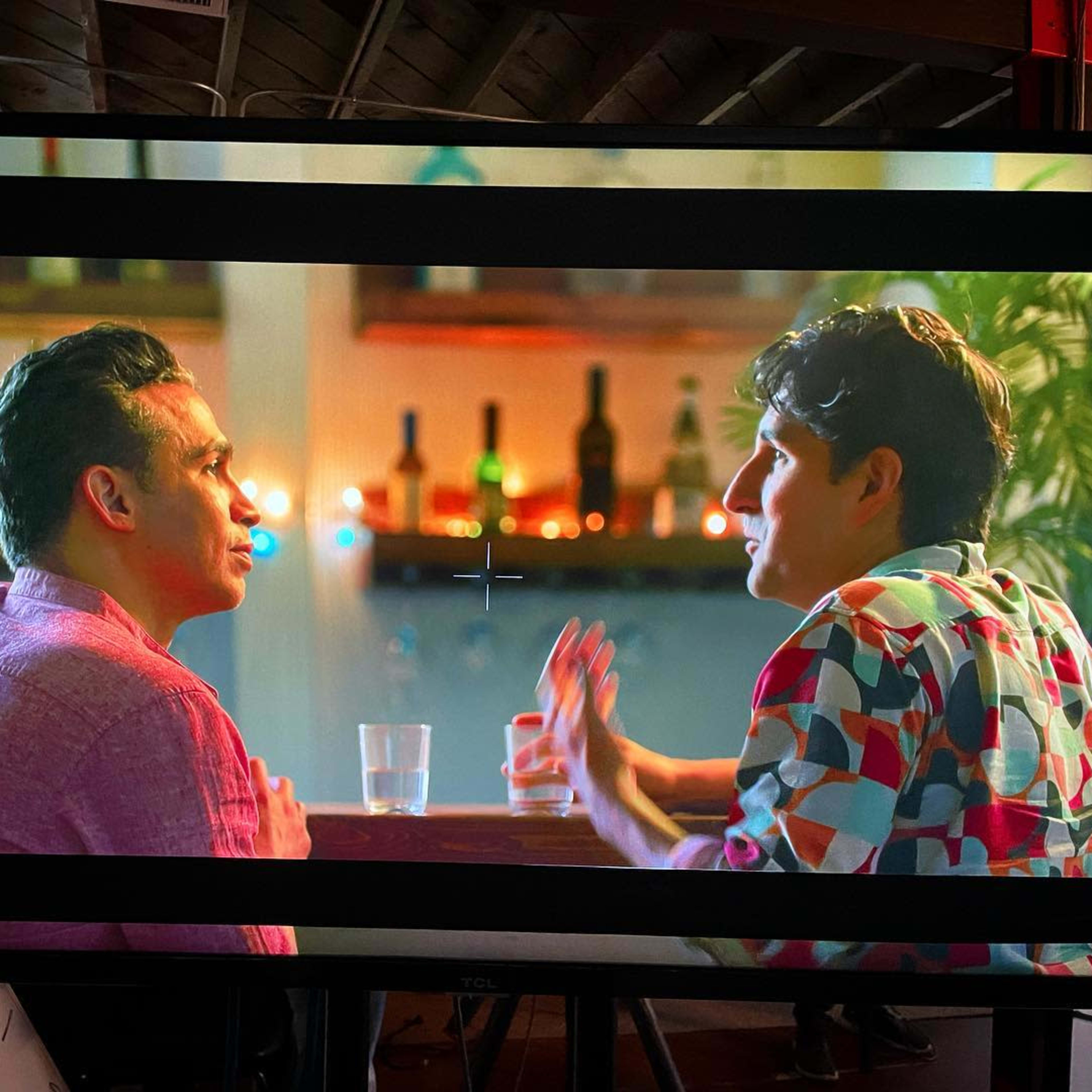 Two men are engaged in a conversation at a bar, with various bottles displayed on shelves in the background.