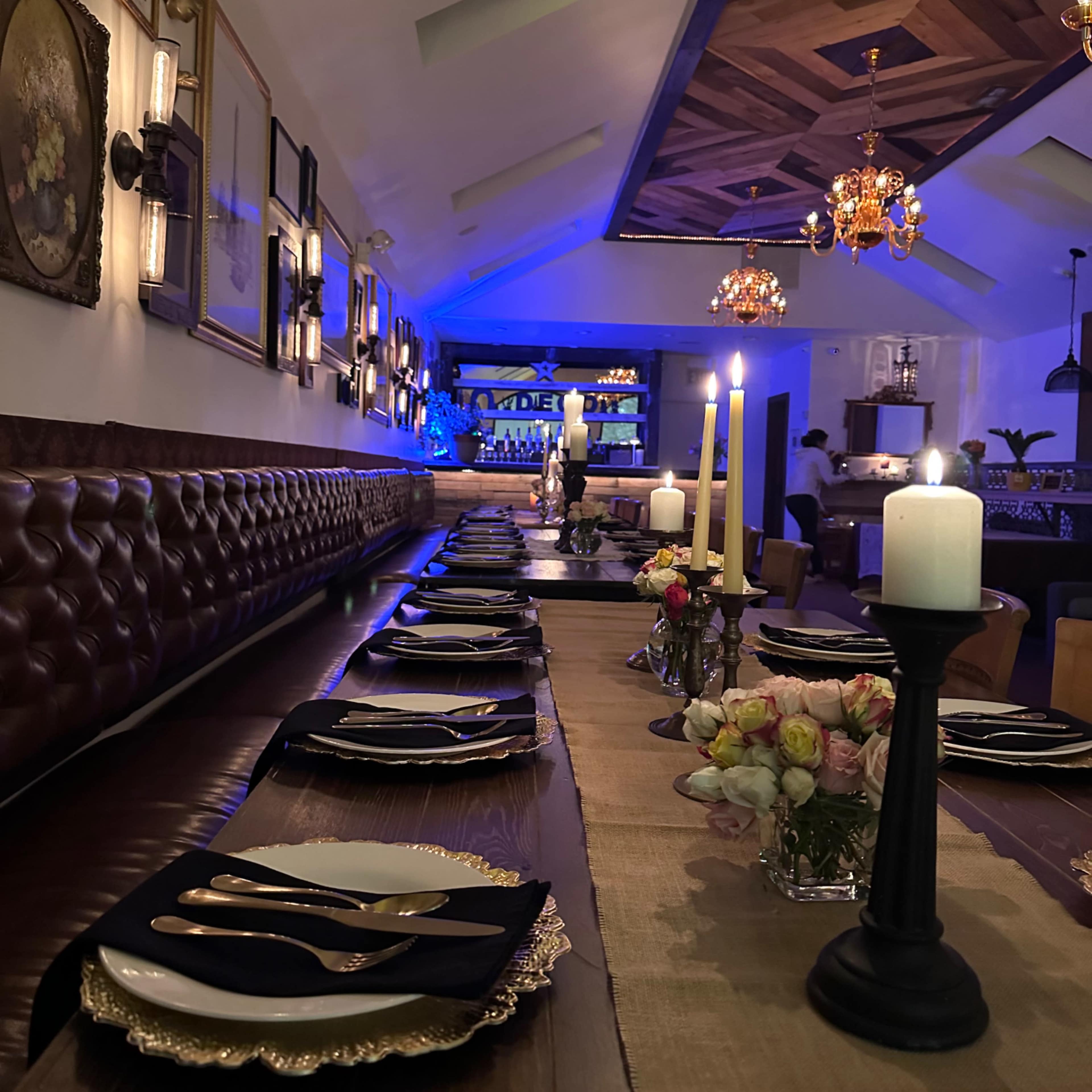 A long dining table set with elegant place settings, candles, and flower arrangements, placed in a restaurant with ambient lighting and decorative wall art.