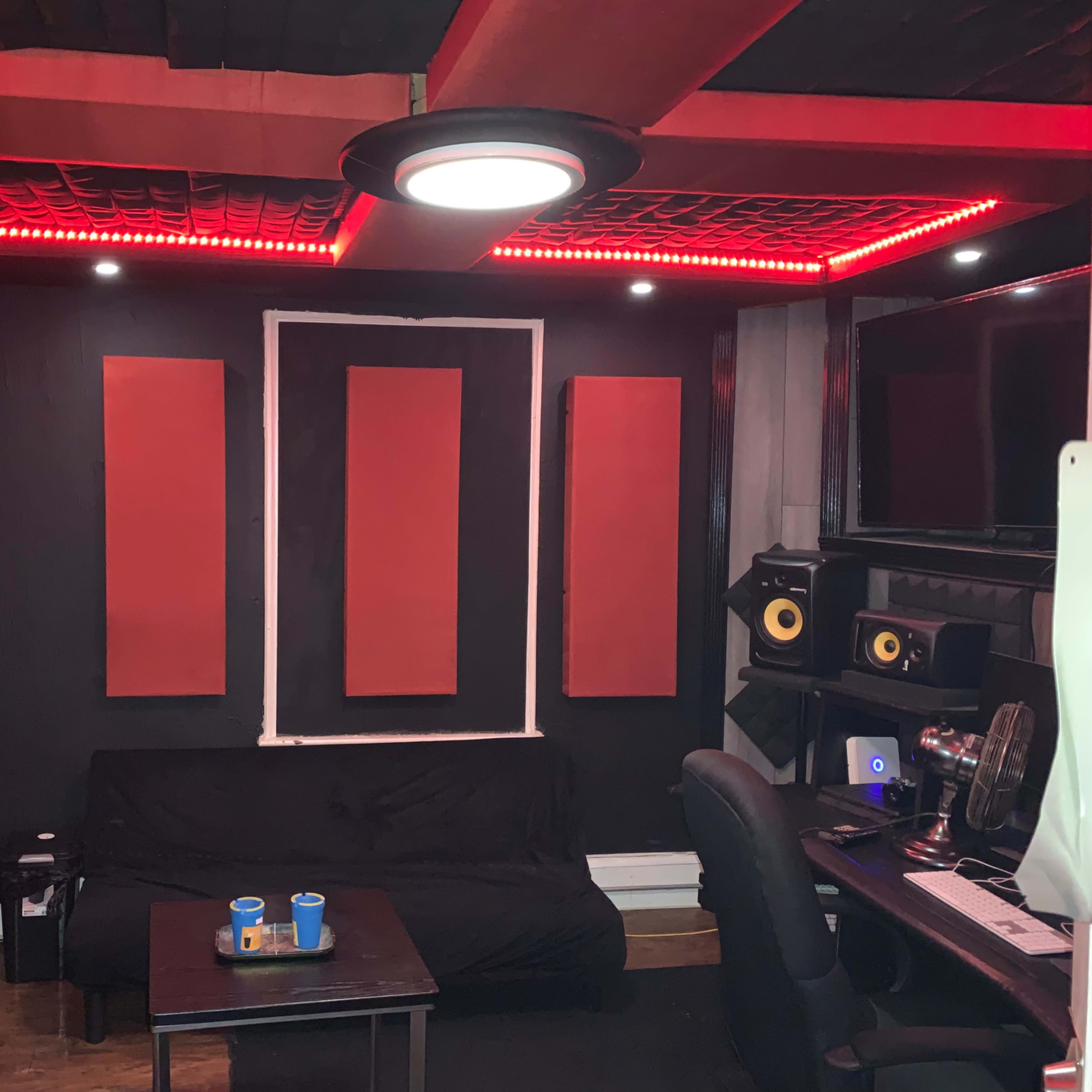 A music studio featuring black walls with red soundproofing panels, a black sofa, a small table with two cups, and audio equipment set up on a desk.