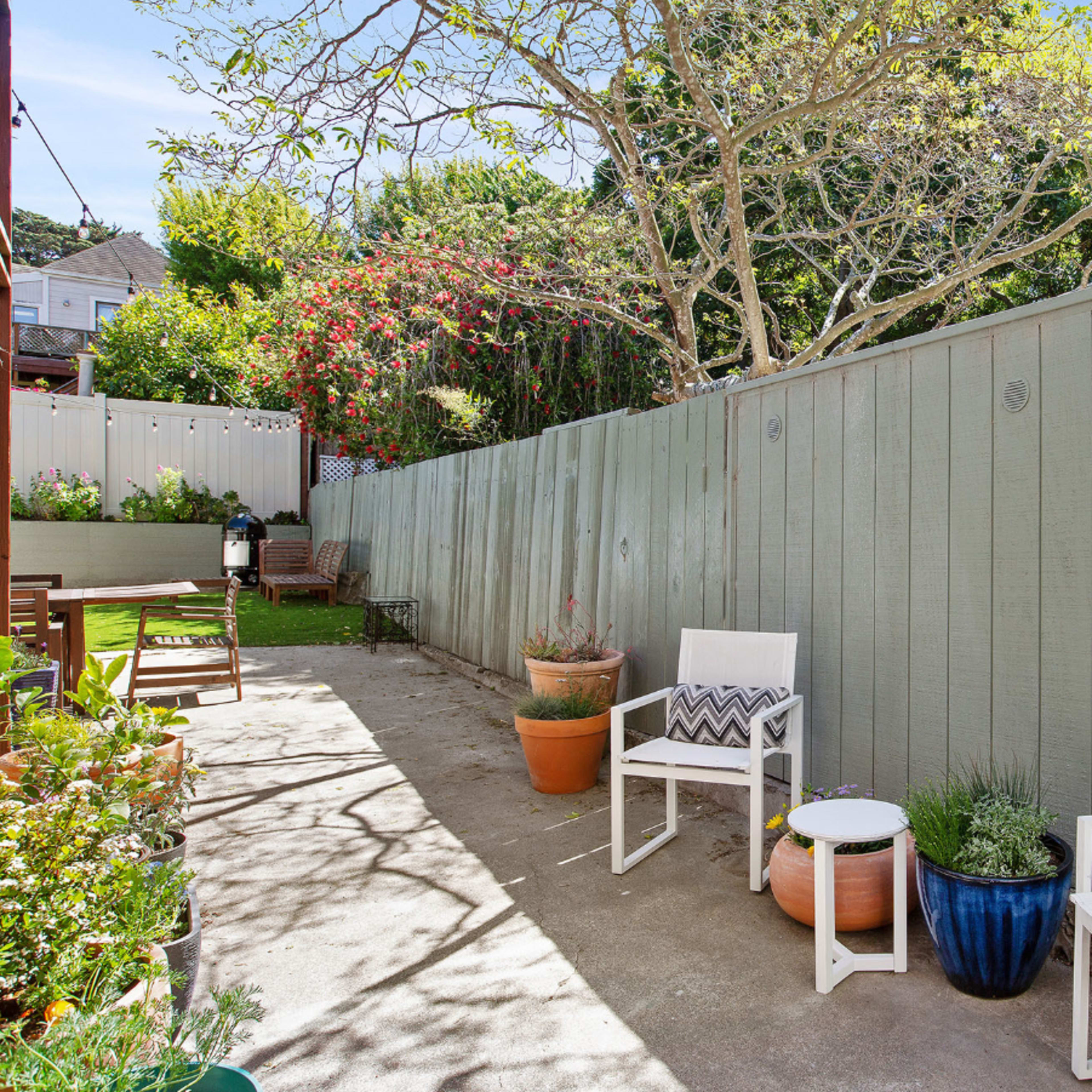 A small outdoor patio area with two white chairs, potted plants, and a wooden fence.