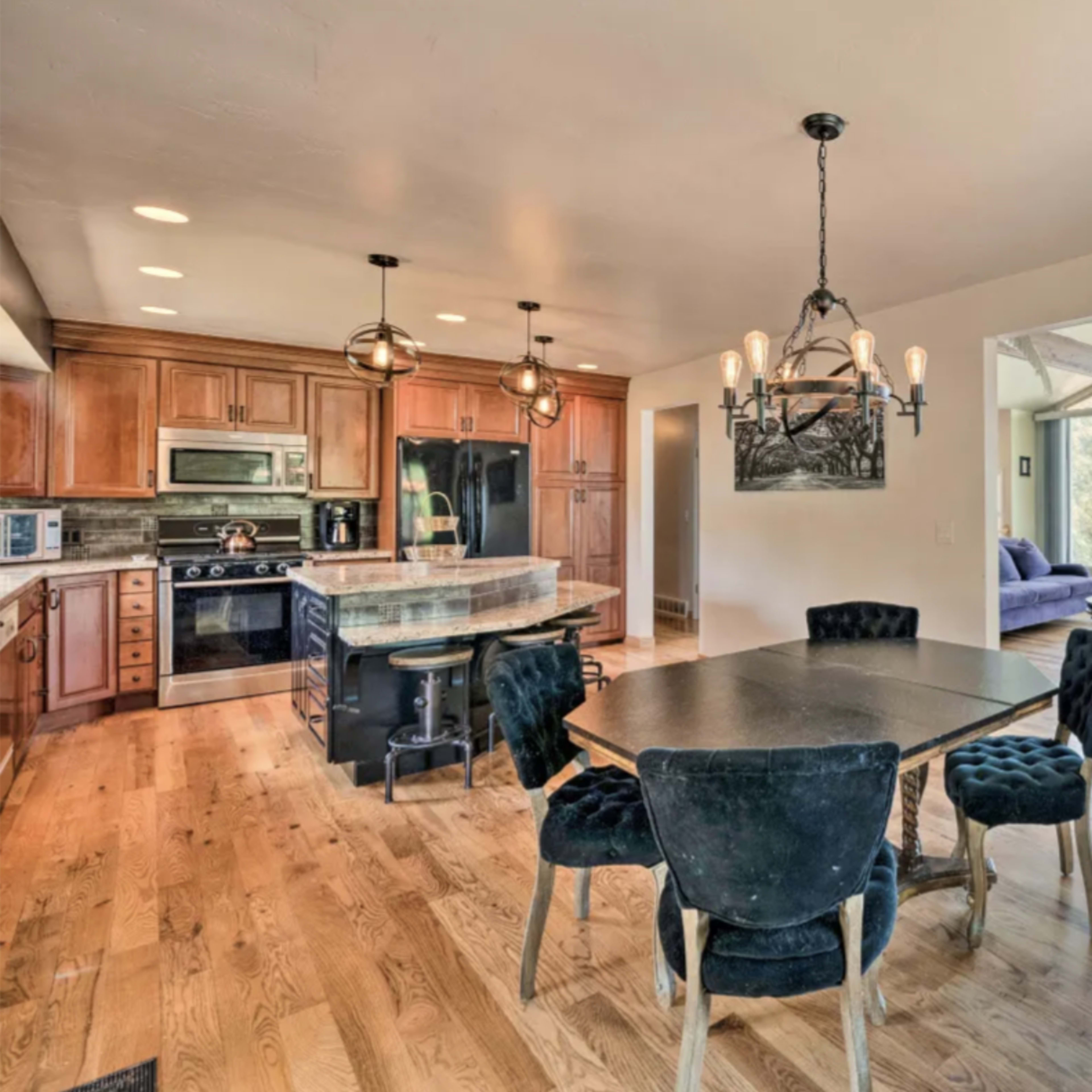 A dining area with a black table and six chairs, adjacent to a kitchen featuring wooden cabinets and modern appliances, with large windows offering a view of the outdoors.