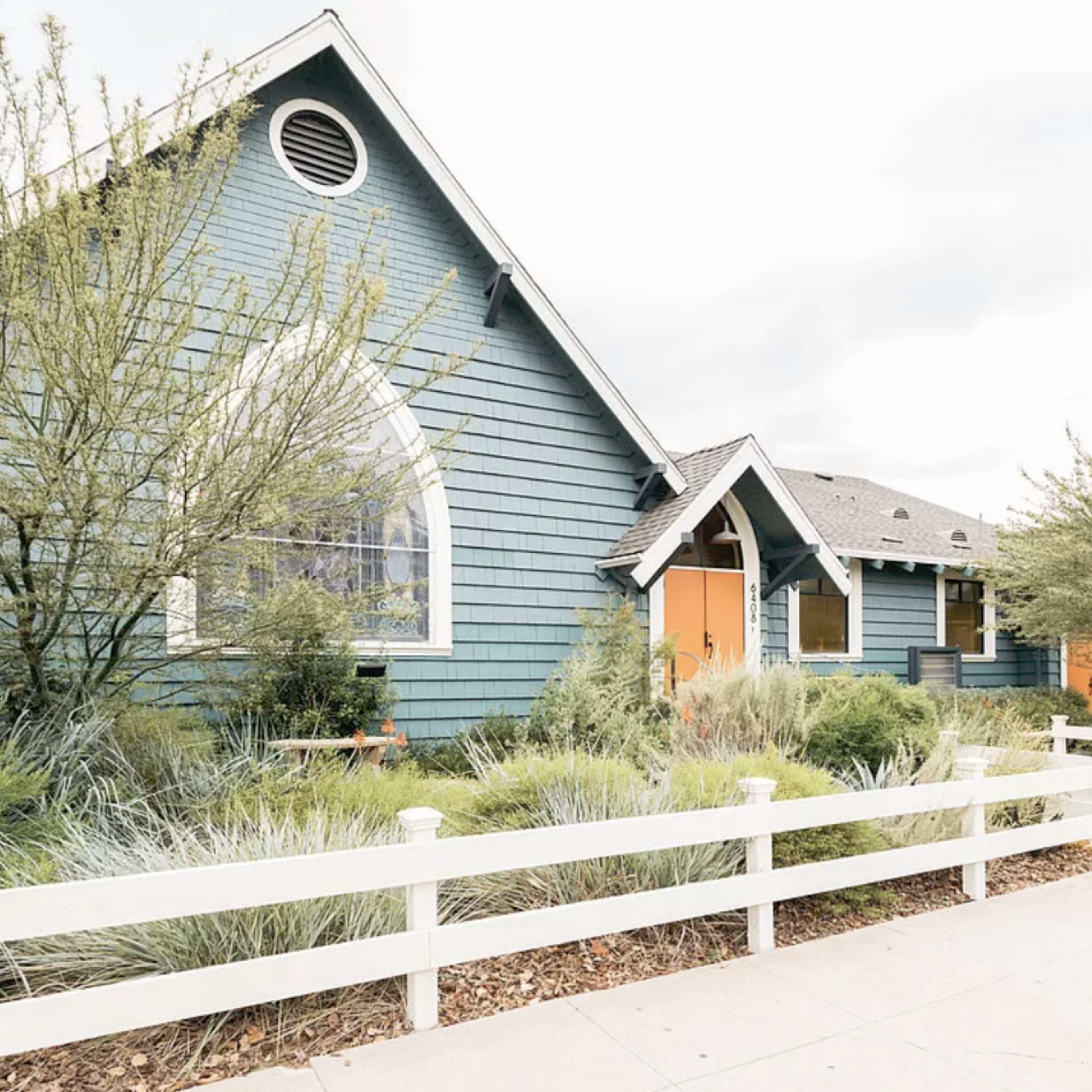 A blue, A-frame house with orange doors is surrounded by greenery and a white picket fence.