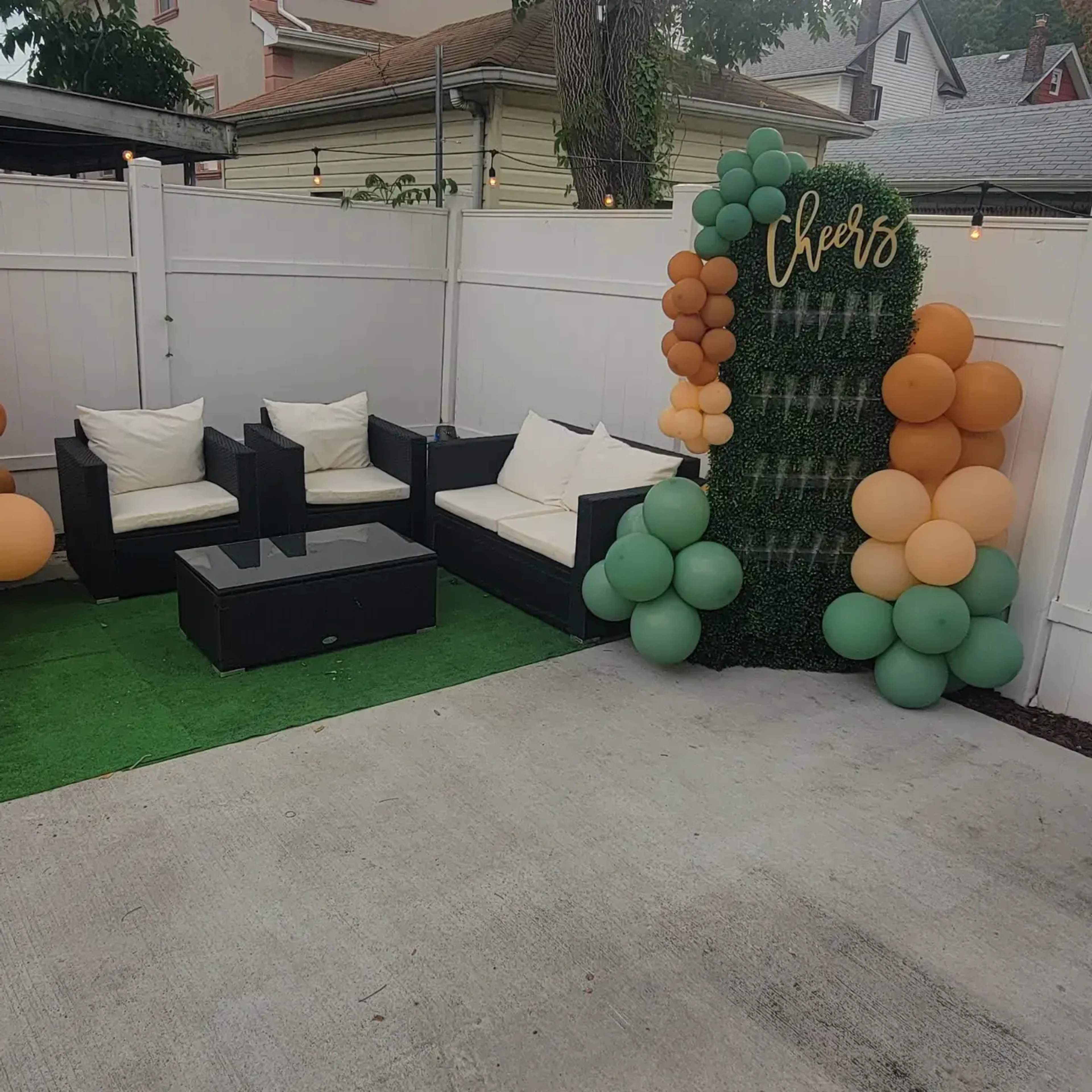 A backyard seating area with black outdoor furniture, a green grass mat, and a decorative balloon arrangement that says "Cheers."