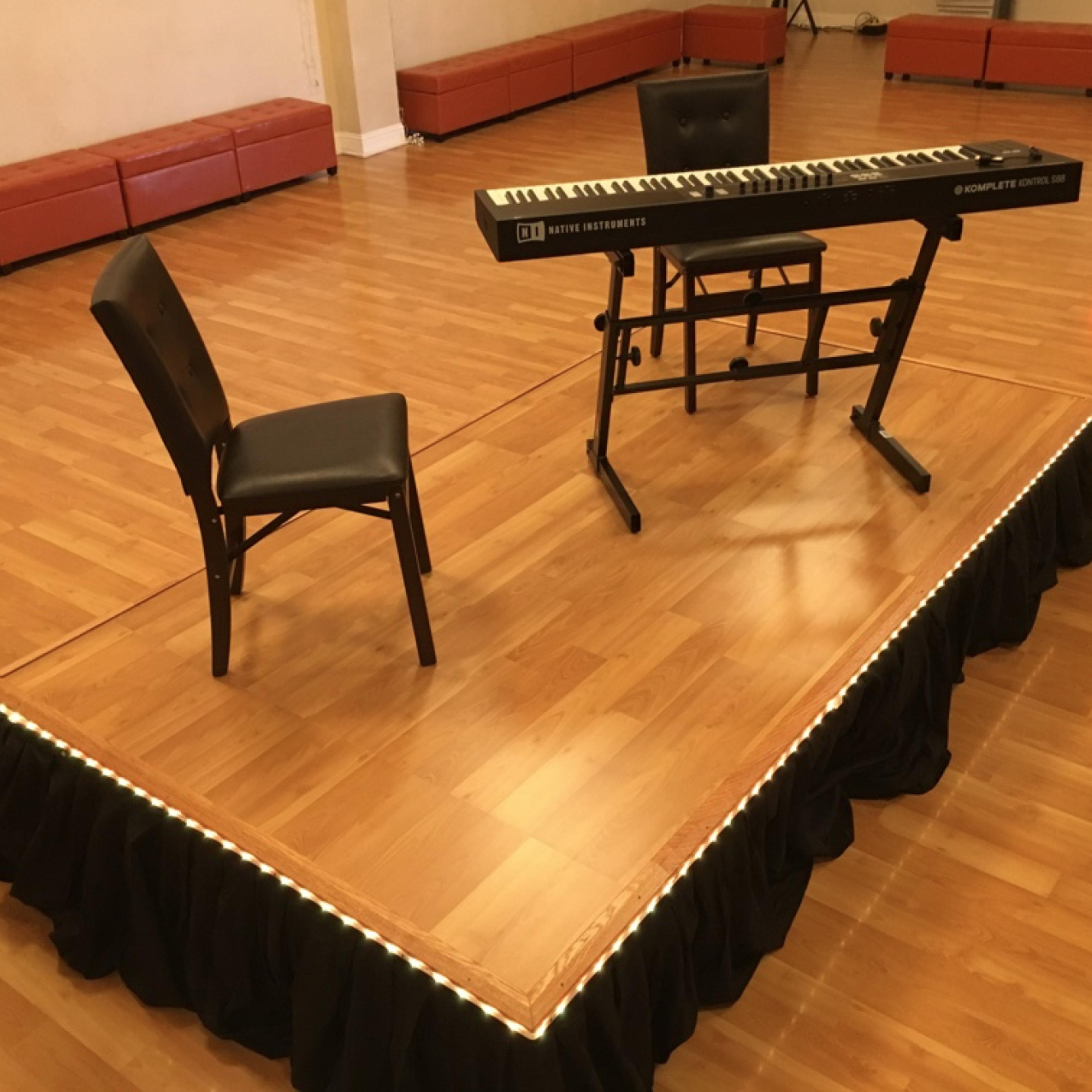 A keyboard and a chair are placed on a small stage with a black draped edge in a wooden-floored room.