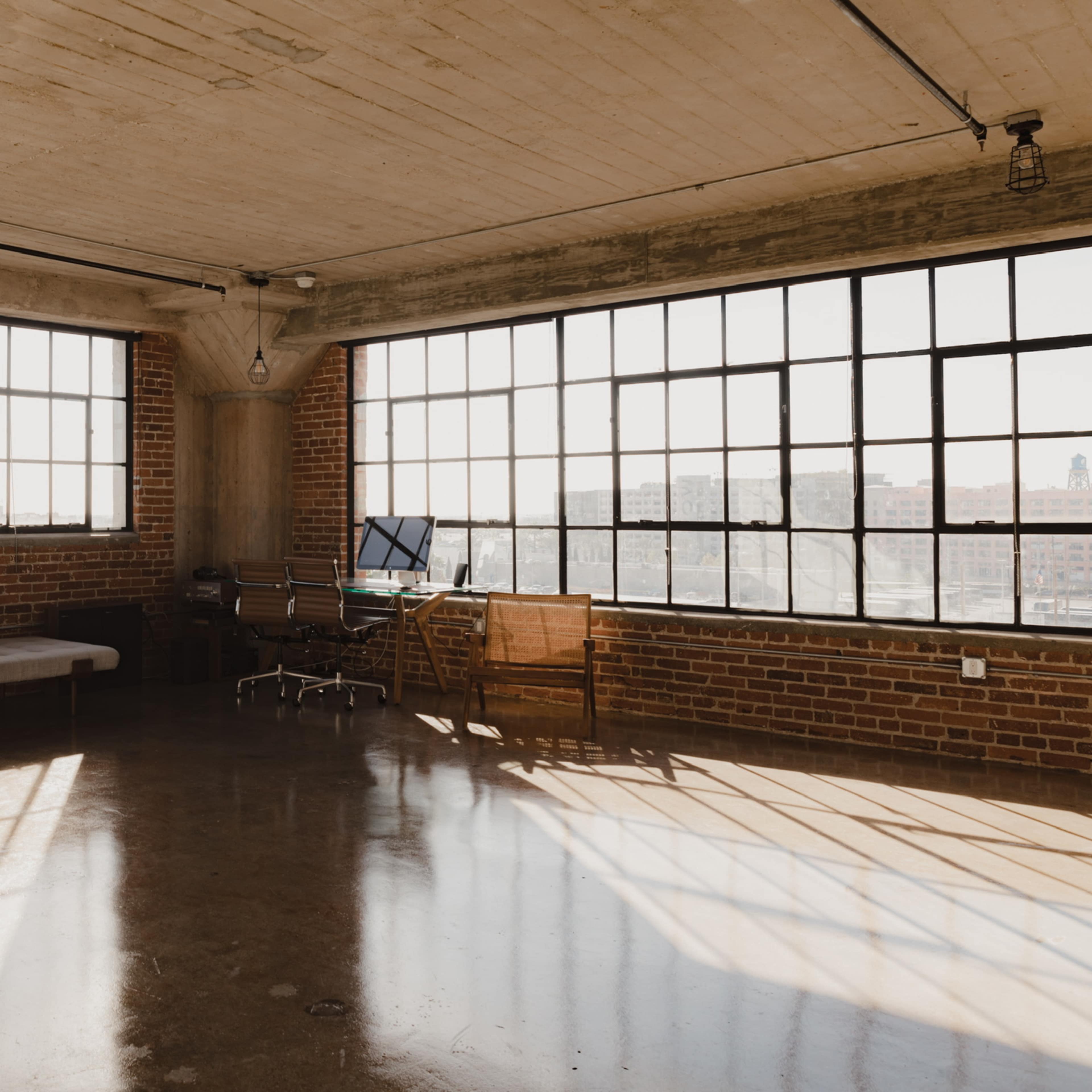 A spacious room with exposed brick walls, large windows, and minimal furniture, including a bed, two chairs, and a desk with a computer.