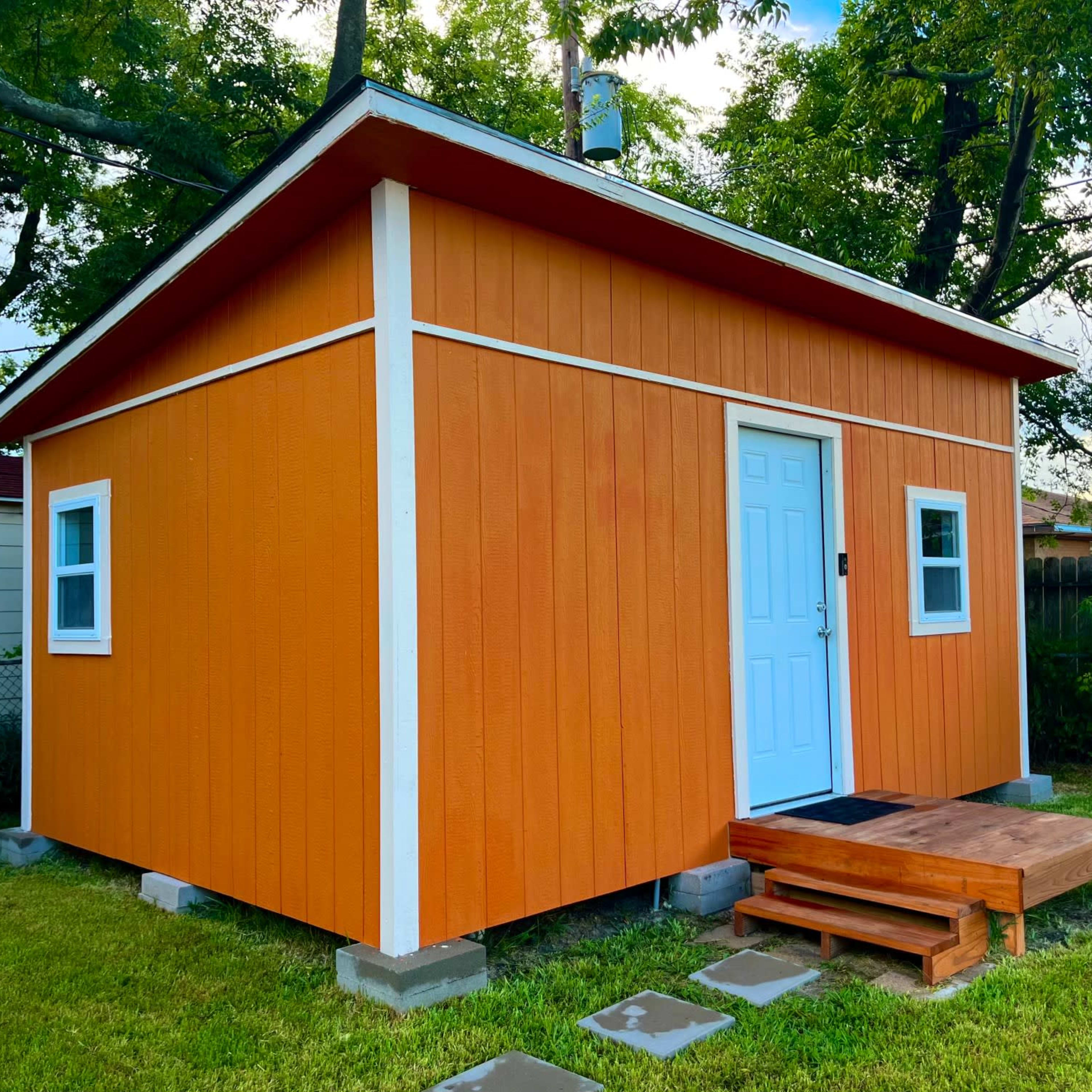 A bright orange shed with a white door and windows is set on a grassy area surrounded by trees and a fence.