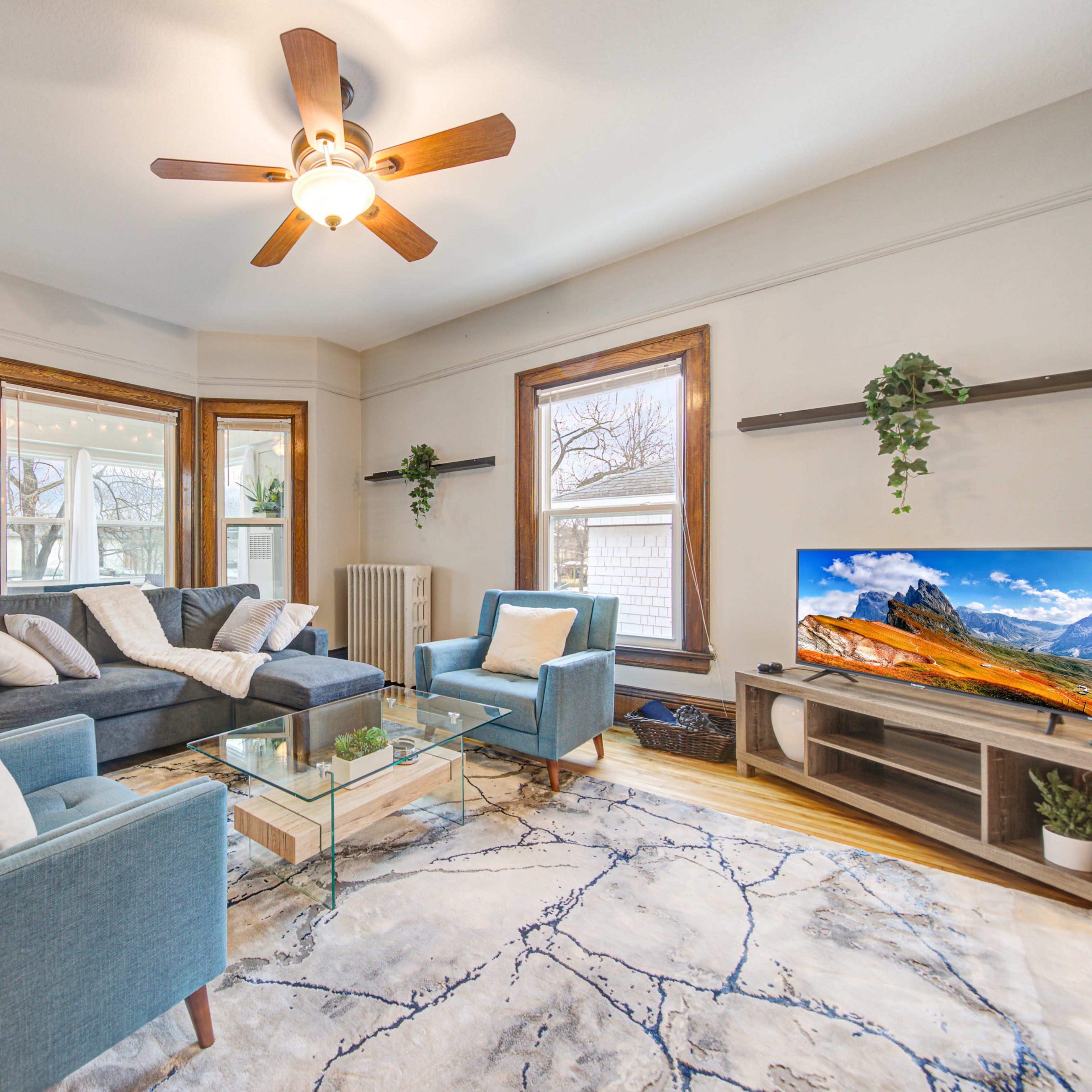 A bright living room features large windows, a ceiling fan, a glass coffee table, and modern furniture with a television displaying a mountain landscape.
