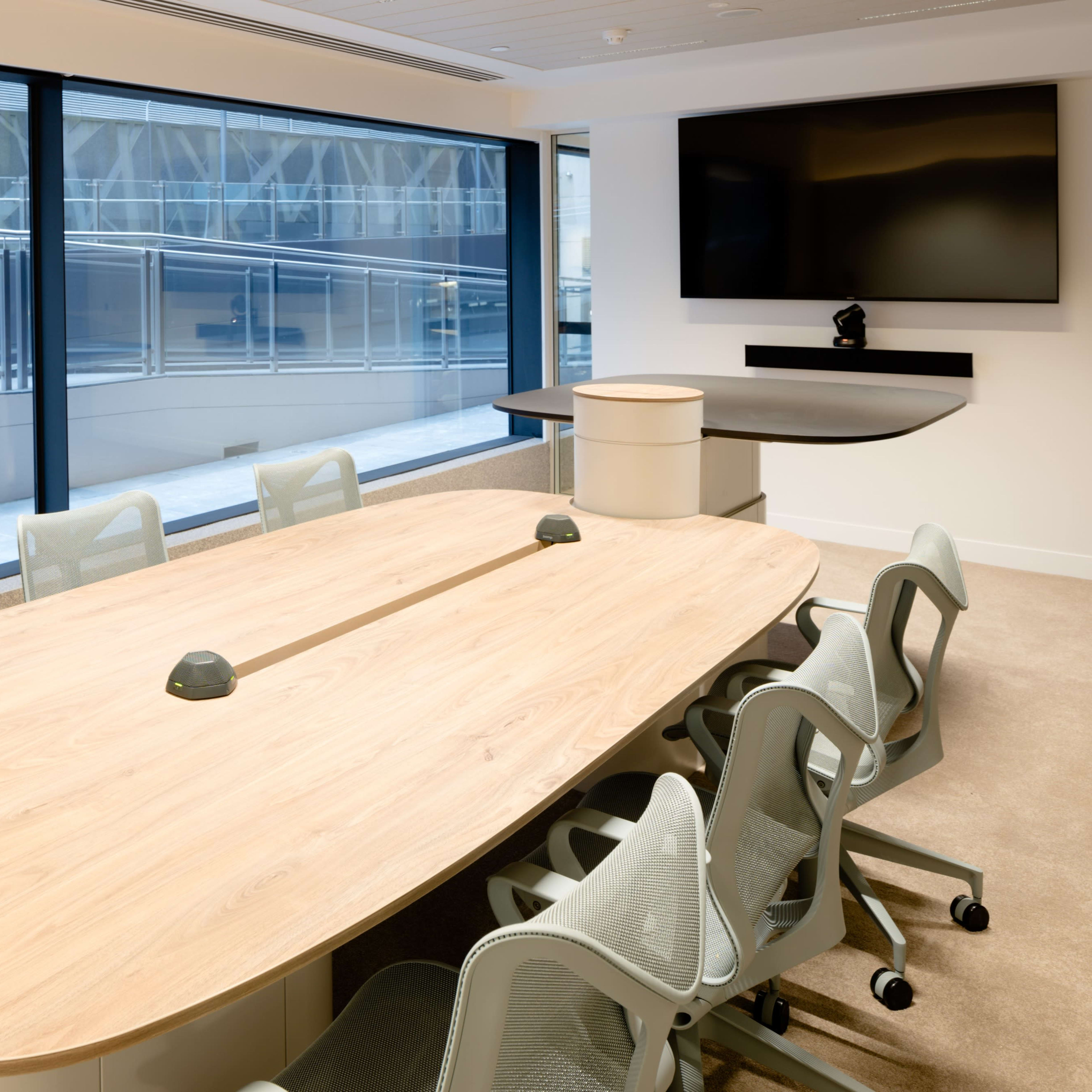 A modern conference room features a large wooden table surrounded by ergonomic chairs and is illuminated by natural light from large windows.