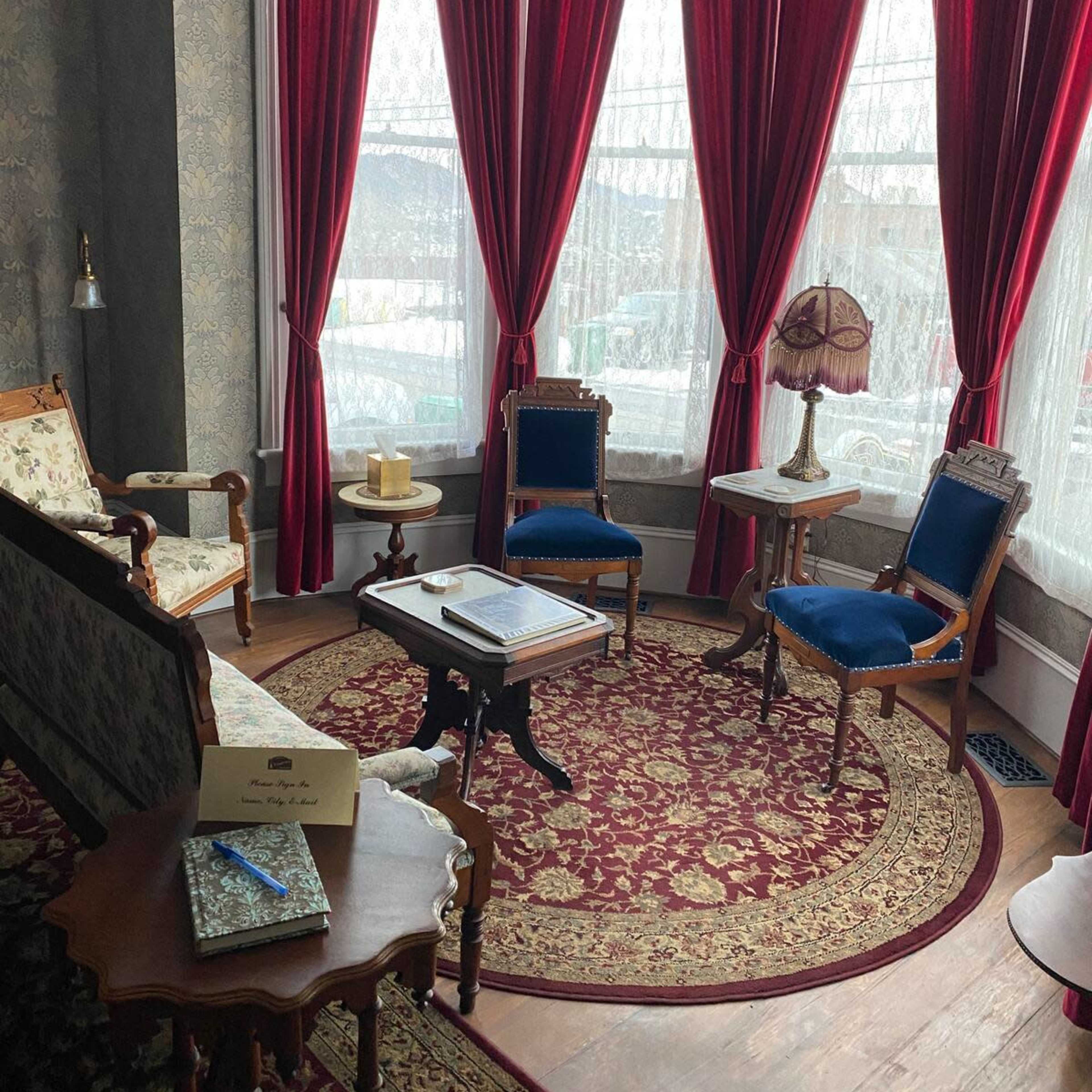 A corner of a room with a patterned rug, vintage furniture, and large red curtains covering a bay window.