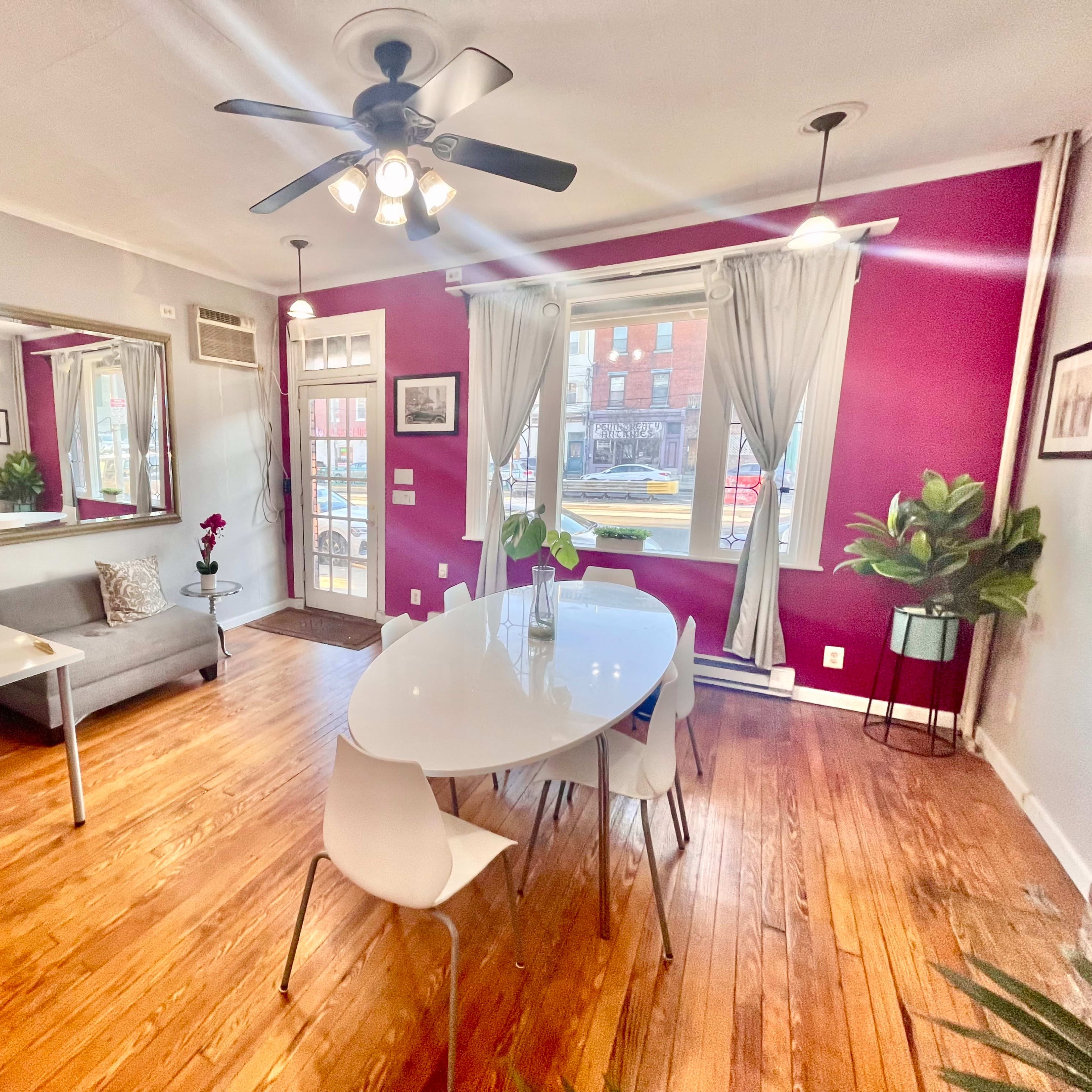 A modern dining area featuring a round table with white chairs, a gray sofa, and a desk, all set against a backdrop of purple and white walls with large windows.