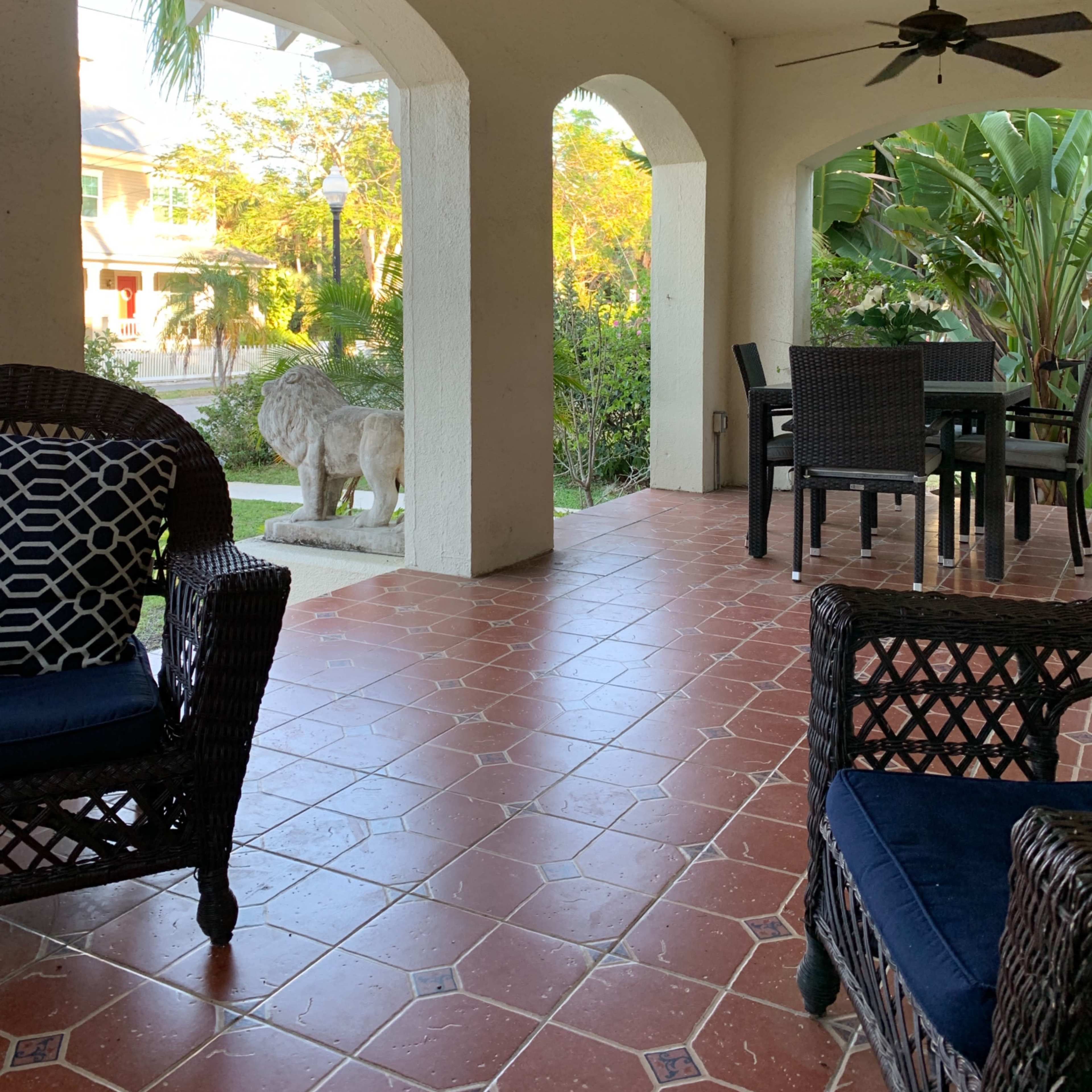 The covered patio features wicker chairs, a table, and a stone lion statue surrounded by tropical plants.