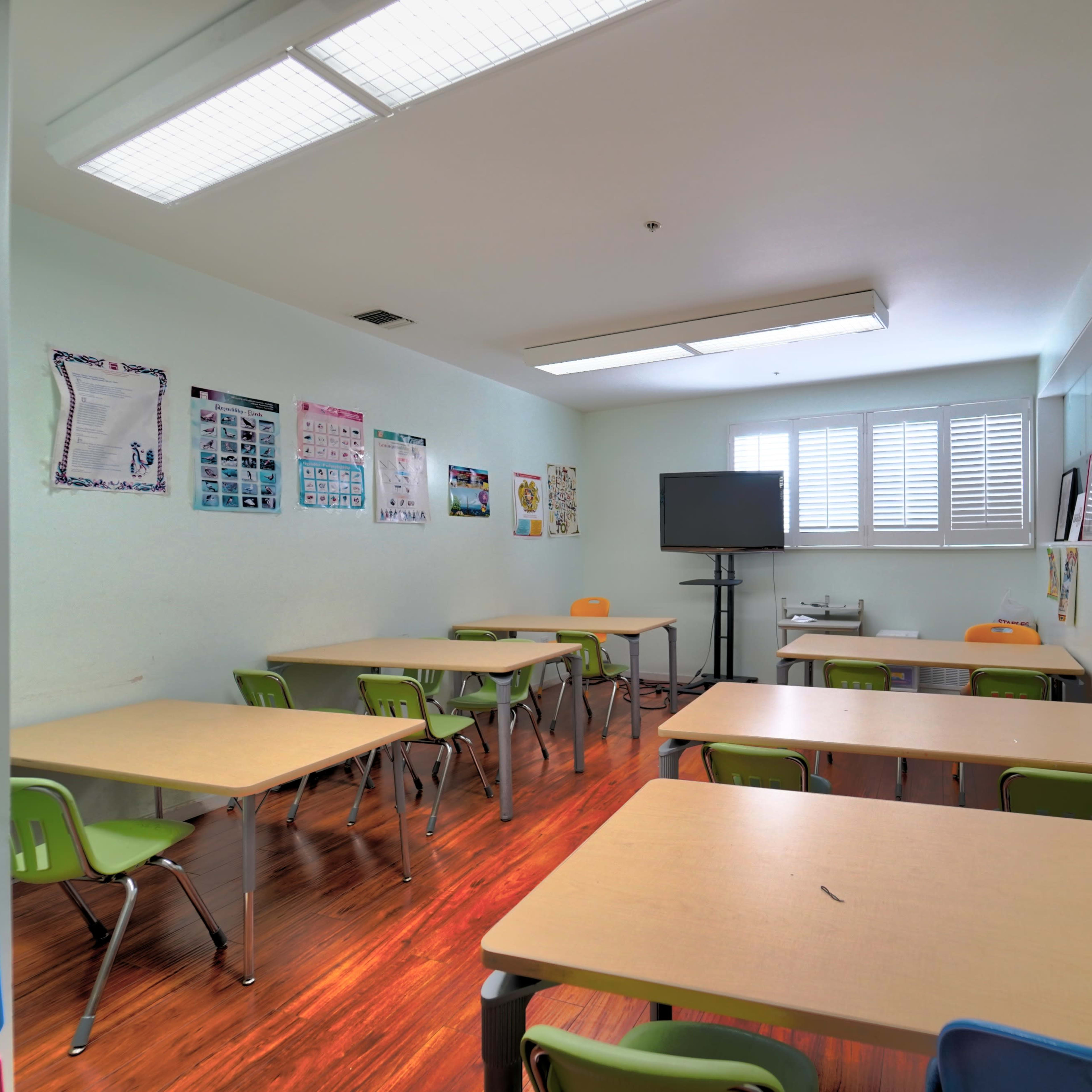 A classroom features several tables with chairs, a large screen, and educational posters on the walls.