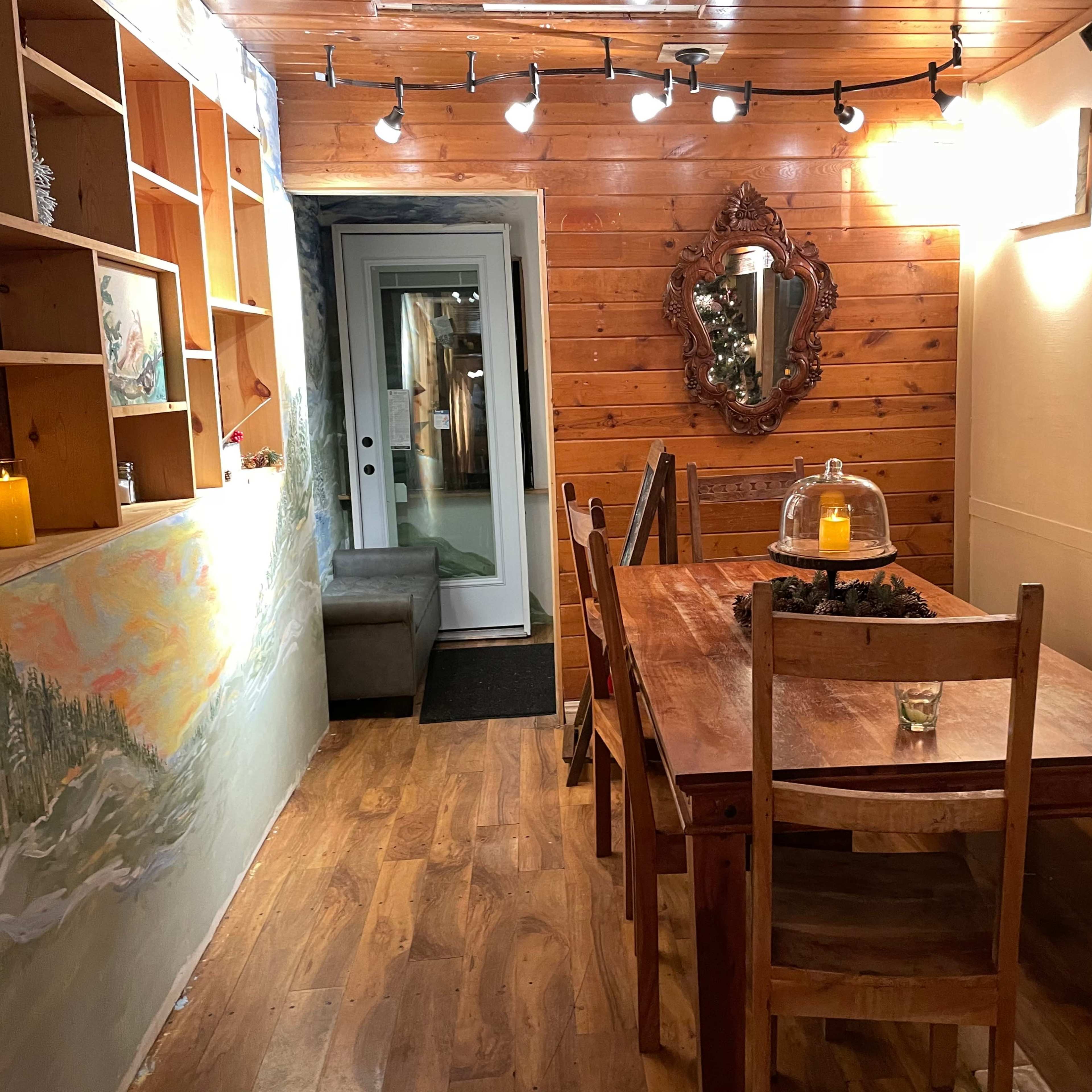 A wooden dining table with chairs in a warmly lit room that features a large mirror and a door leading to an outdoor area.