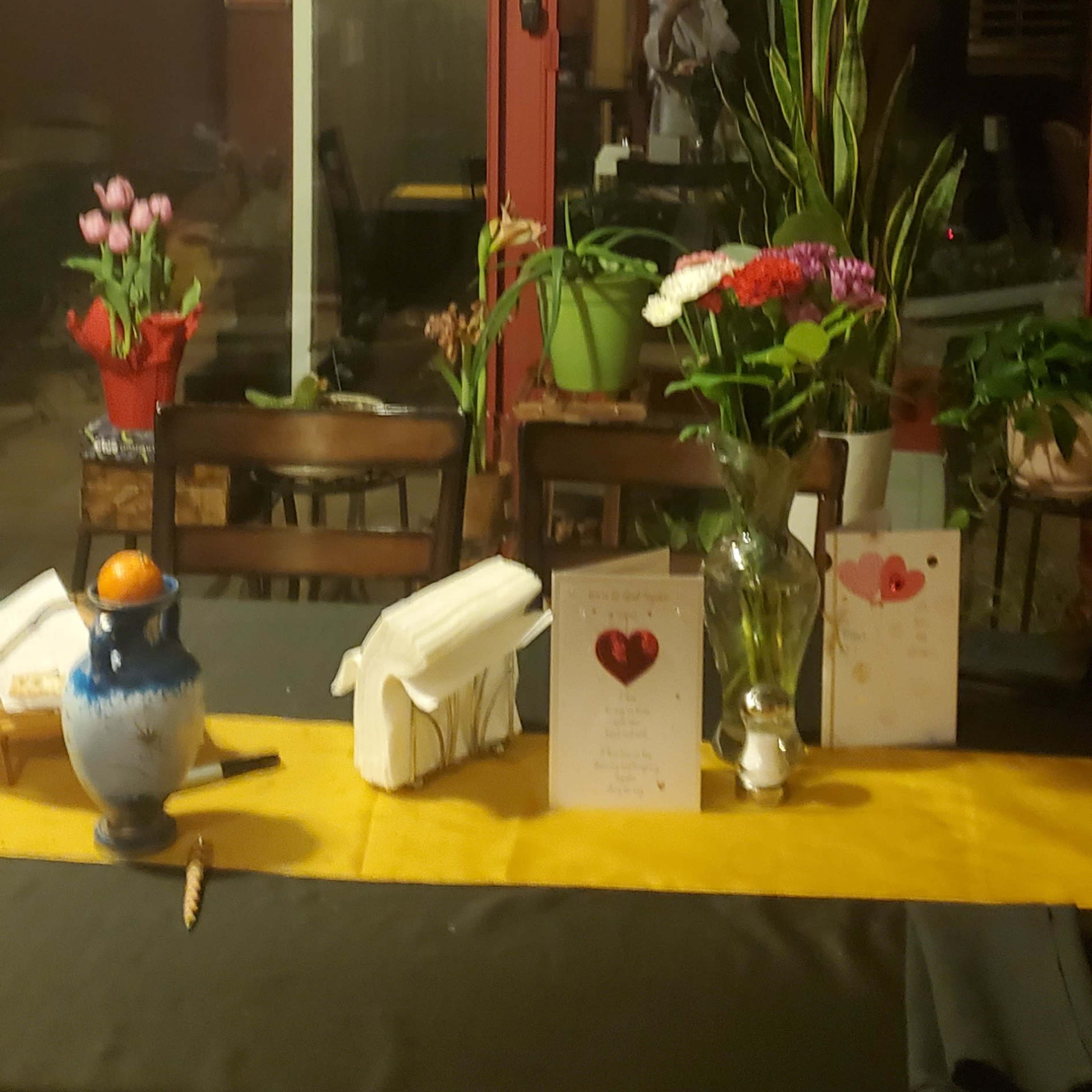 A dining table set with a black tablecloth, a yellow runner, decorative flowers, and greeting cards, with a reflection of a person in the background.