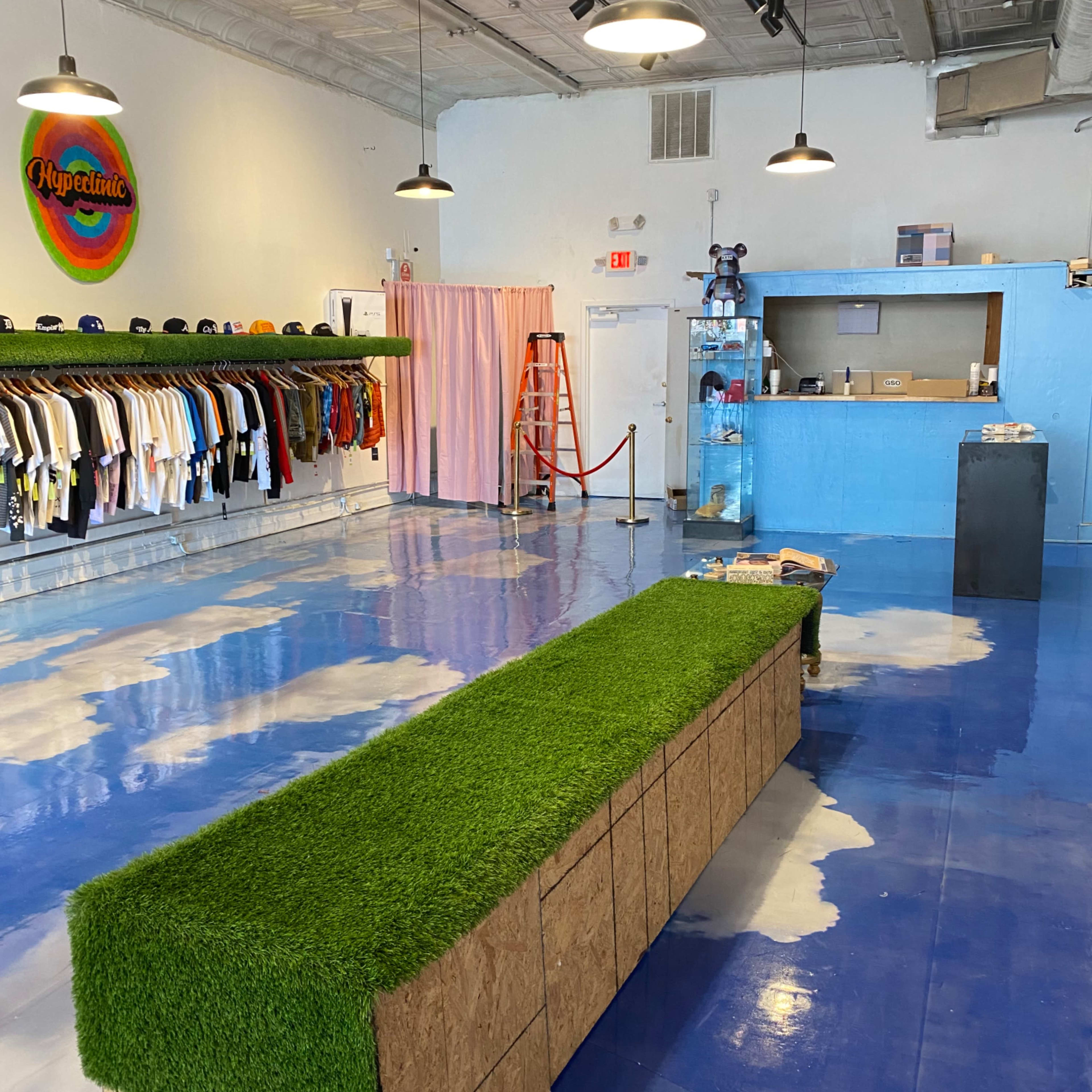 A retail space with clothing displayed on racks, a long green grass-like bench in the center, and a blue counter at the back.